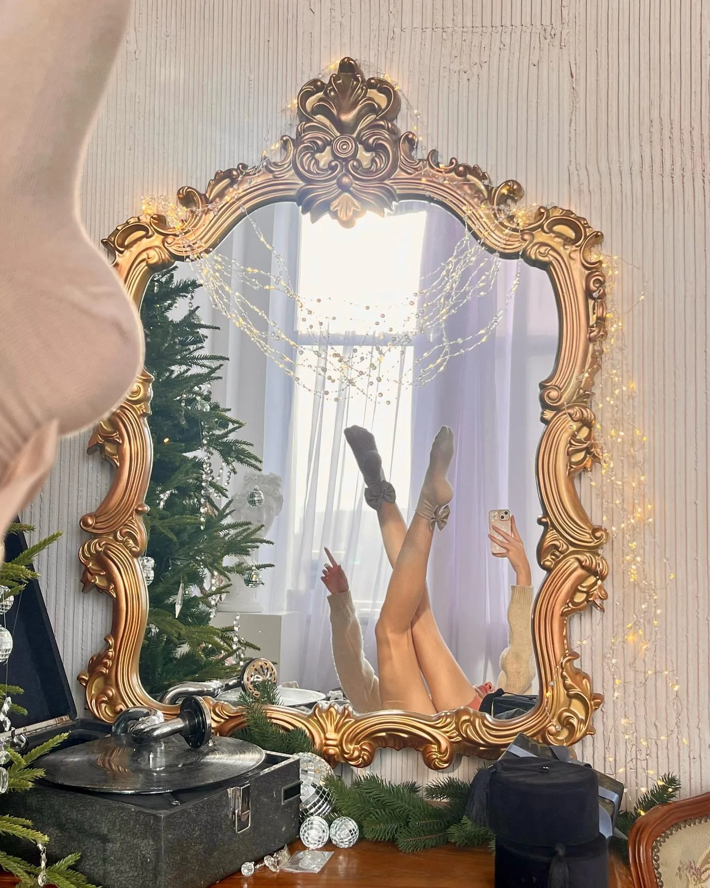 Reflection of a person lying on their back in an ornate gold-framed mirror, with legs raised and holding a phone, next to a Christmas tree.
