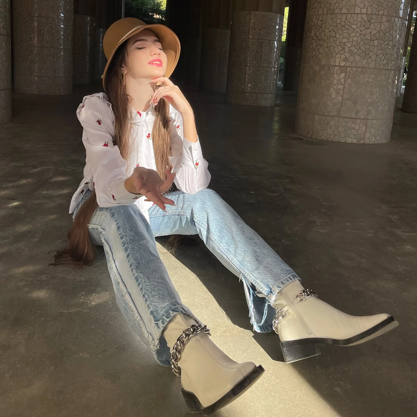 A woman wearing a straw hat, white blouse, and jeans sits on the floor in a sunlit area with large pillars.