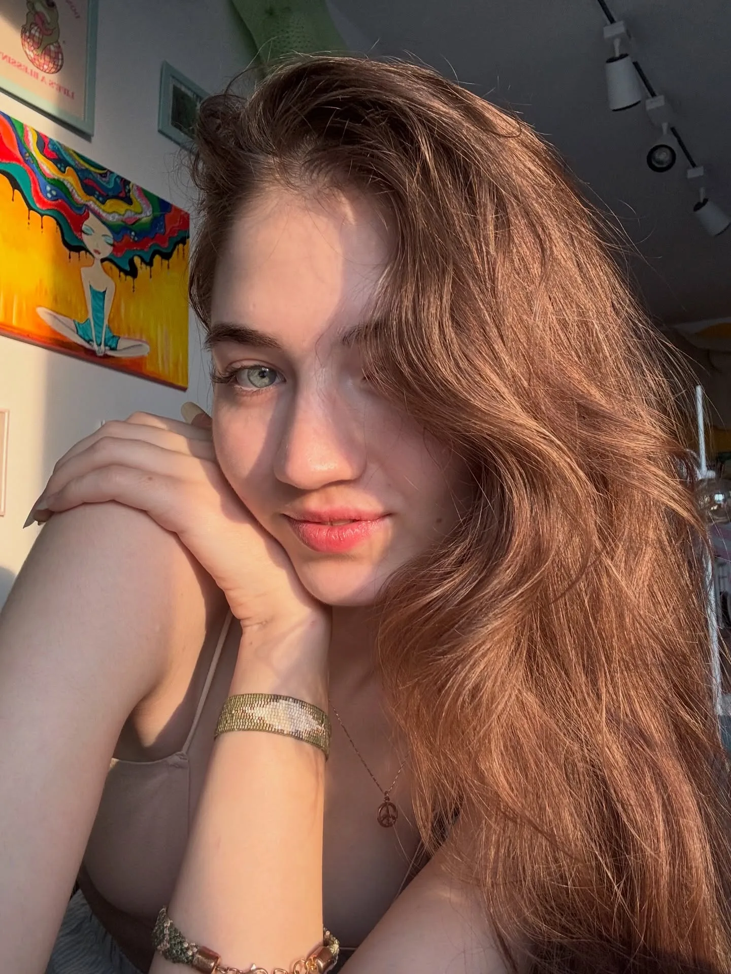 Portrait of a young woman with long wavy brown hair resting her chin on her hand, wearing a gold bracelet and a grey top, with colorful artwork visible in the background.