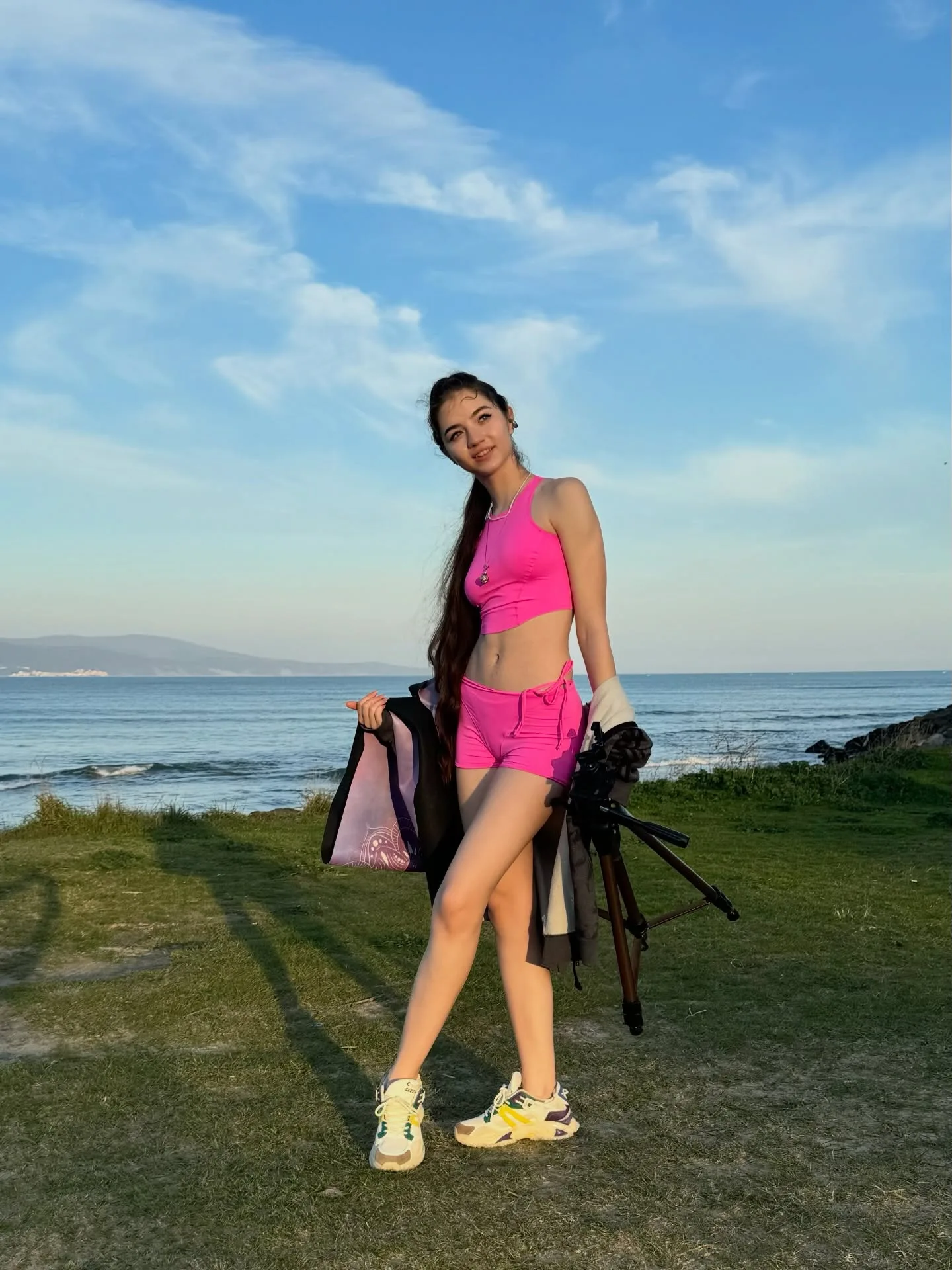 A woman in a pink athletic outfit stands on a grassy hill overlooking the ocean, holding a tripod and a bag under a blue sky.