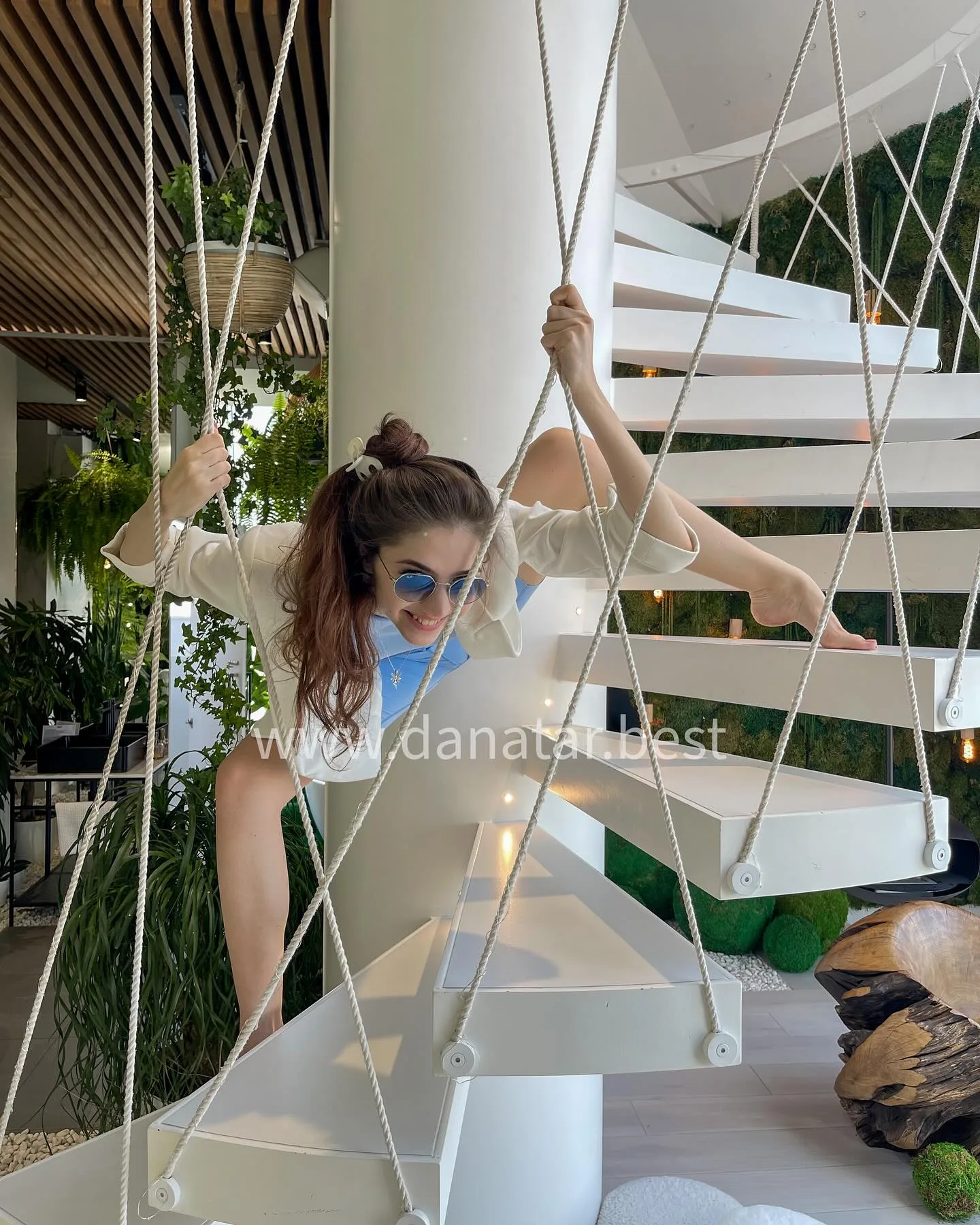 Woman in sunglasses and face mask climbing a modern white spiral staircase with rope railings.