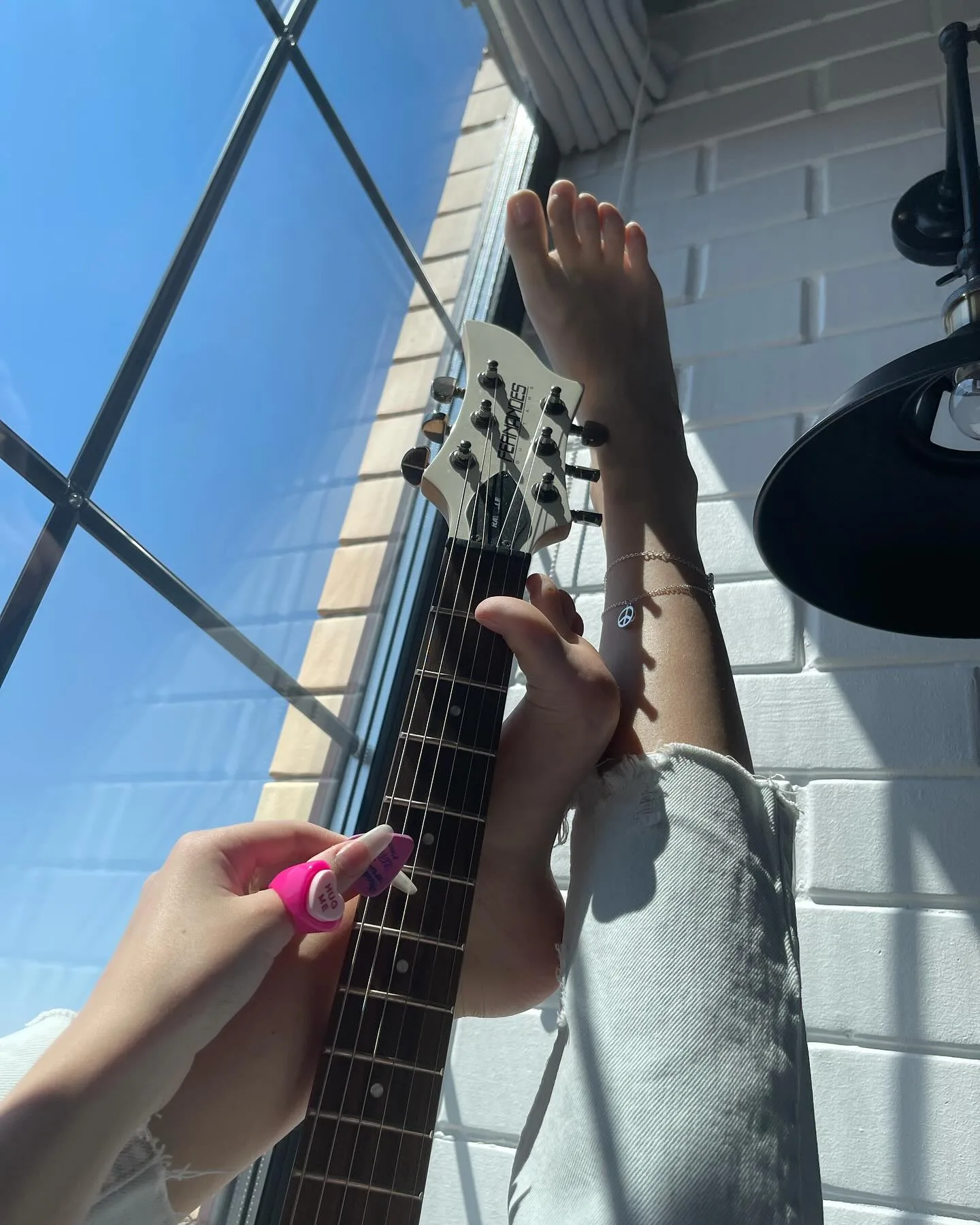 A person playing an acoustic guitar with their feet while holding a pink nail polish bottle in their hand against a bright blue sky.