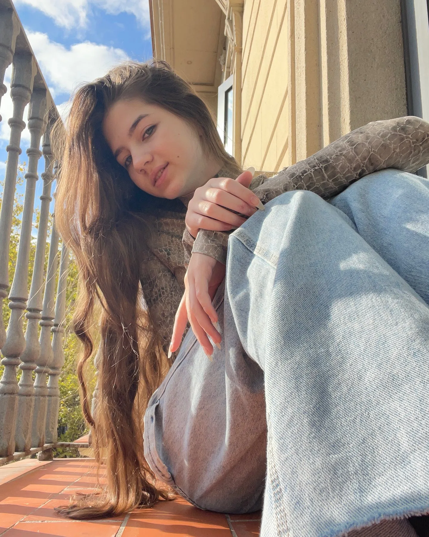 Dana sitting on a tiled balcony with long wavy hair, wearing a brown sweater and light blue jeans, resting her head on her knees against a yellow wall.