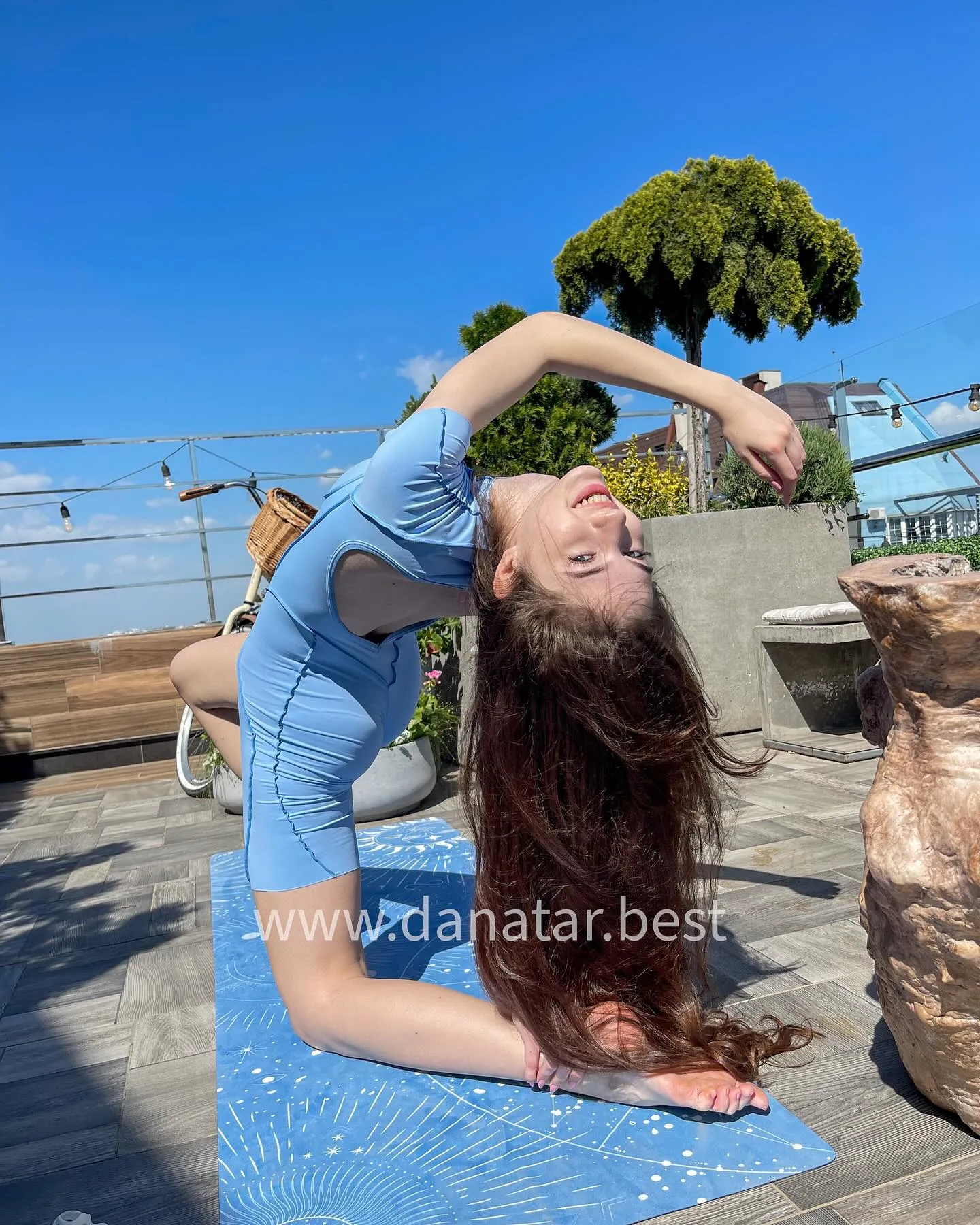 Woman in blue bodysuit performing a deep backbend yoga pose on a blue mat on a sunny rooftop terrace.