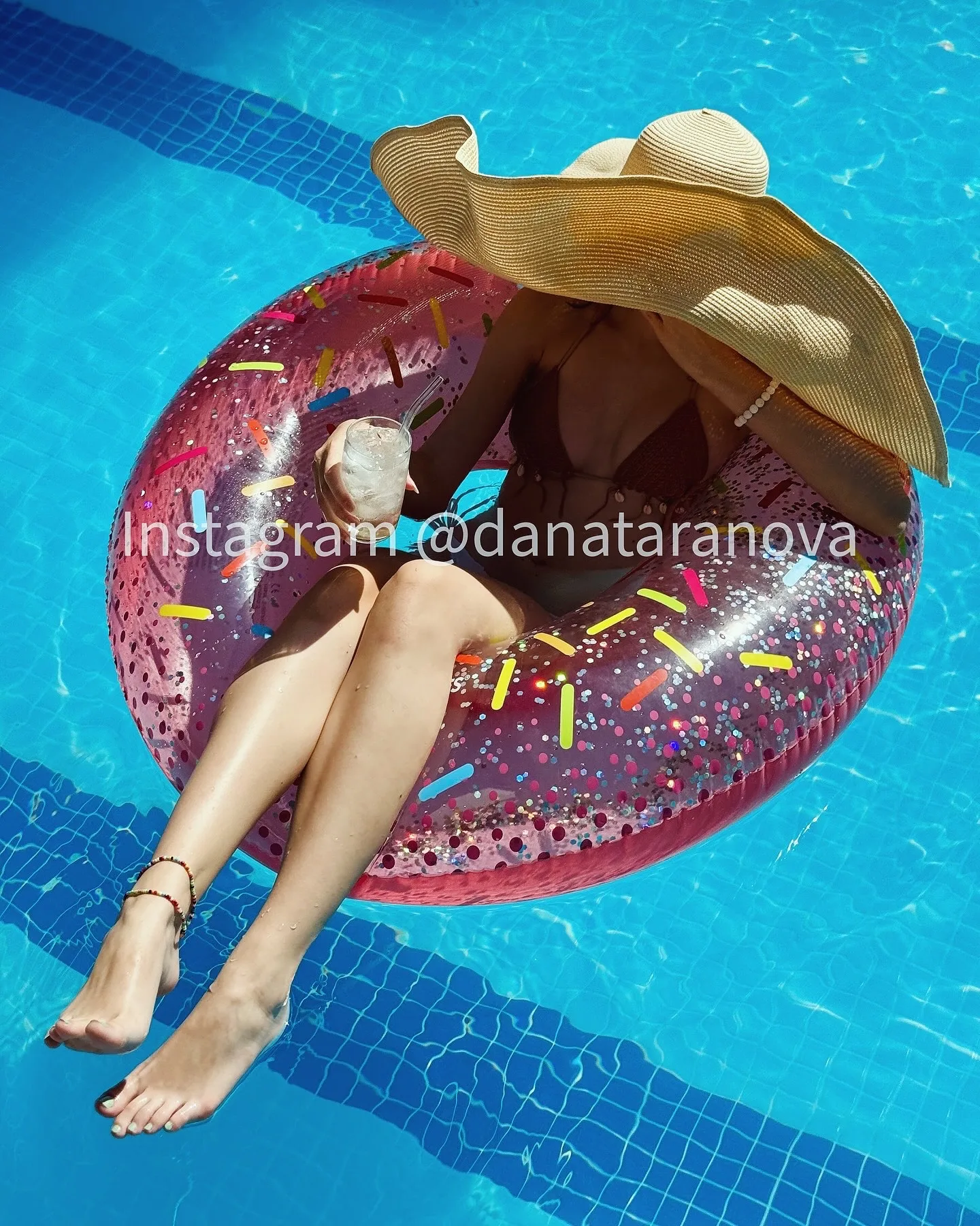 A woman wearing a large straw hat and bikini relaxes on a pink donut-shaped inflatable ring in a blue swimming pool.