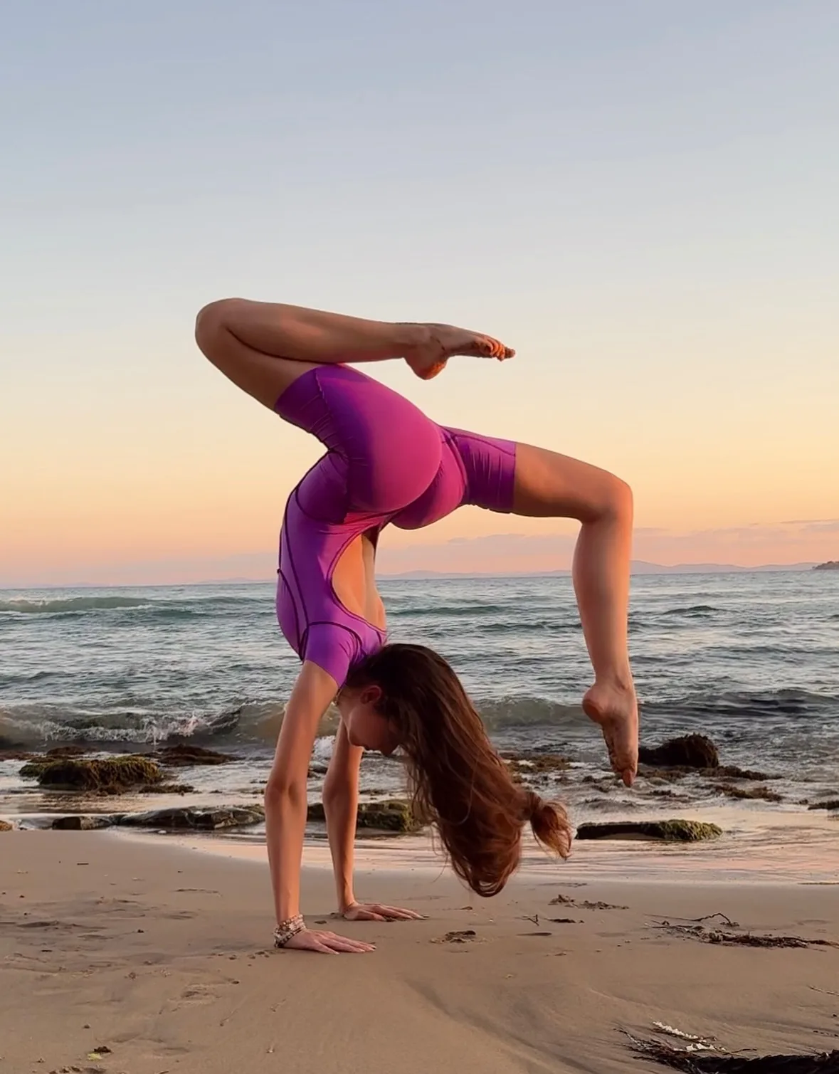 Woman in purple athletic wear performing a handstand on a sandy beach at sunset with ocean waves in the background.