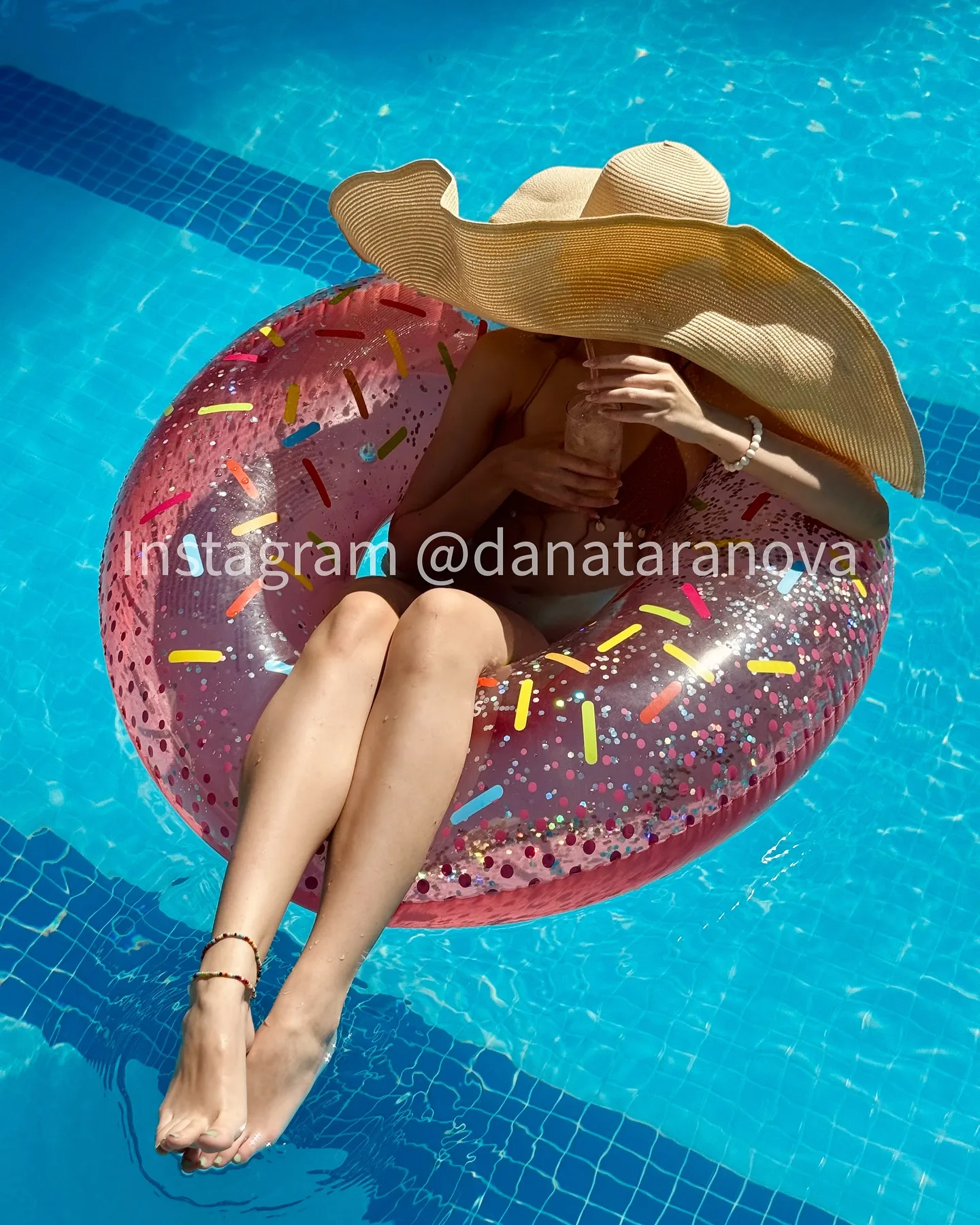 Woman in a wide-brimmed hat relaxing on a pink donut-shaped inflatable ring in a blue swimming pool.