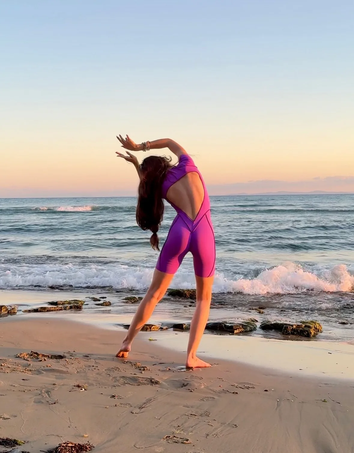 A woman in a purple swimsuit poses on a sandy beach at sunset with arms raised and ocean waves in the background.