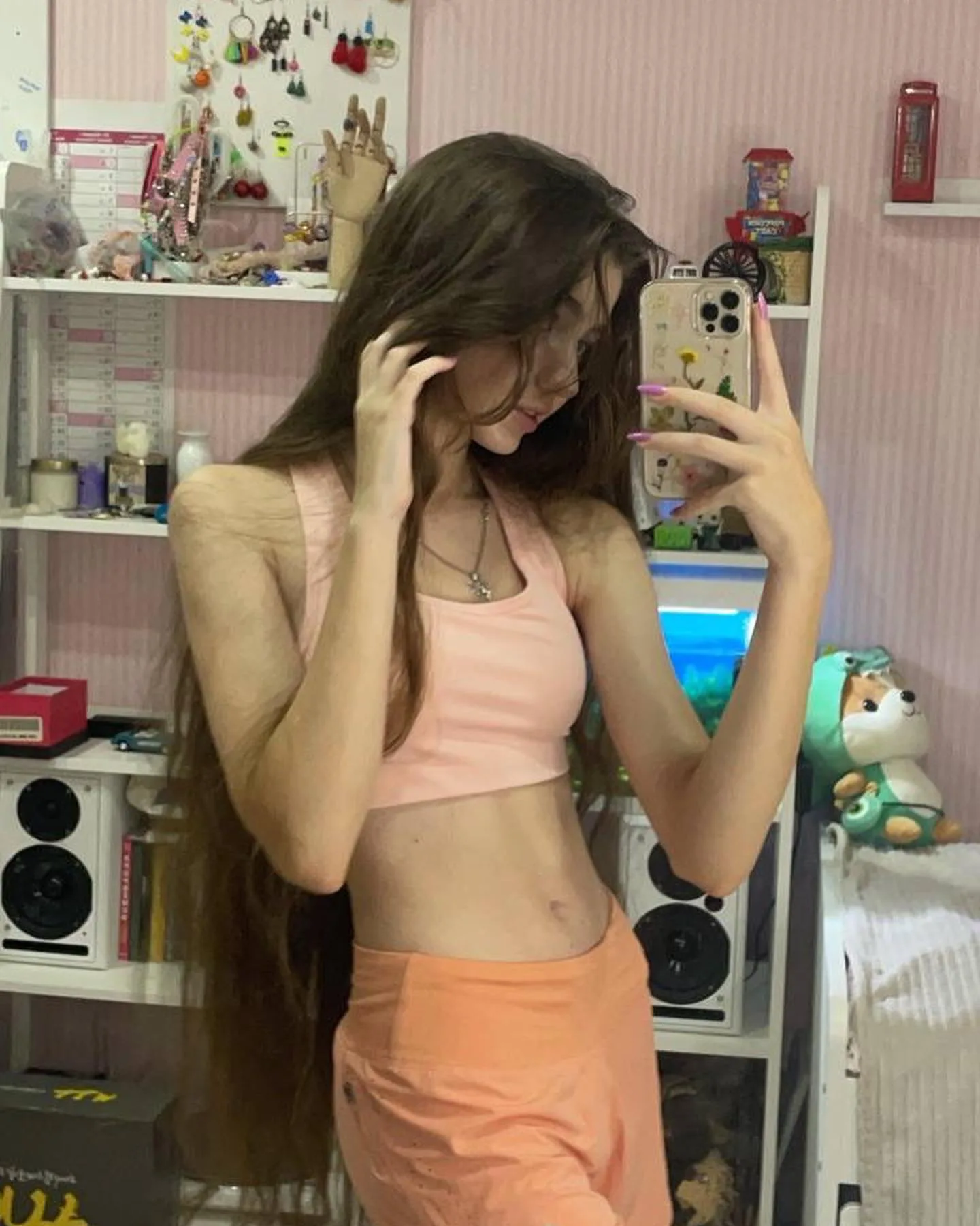 A young woman with long brown hair takes a mirror selfie in a pink top and orange skirt, standing in a room with white shelves filled with various items.