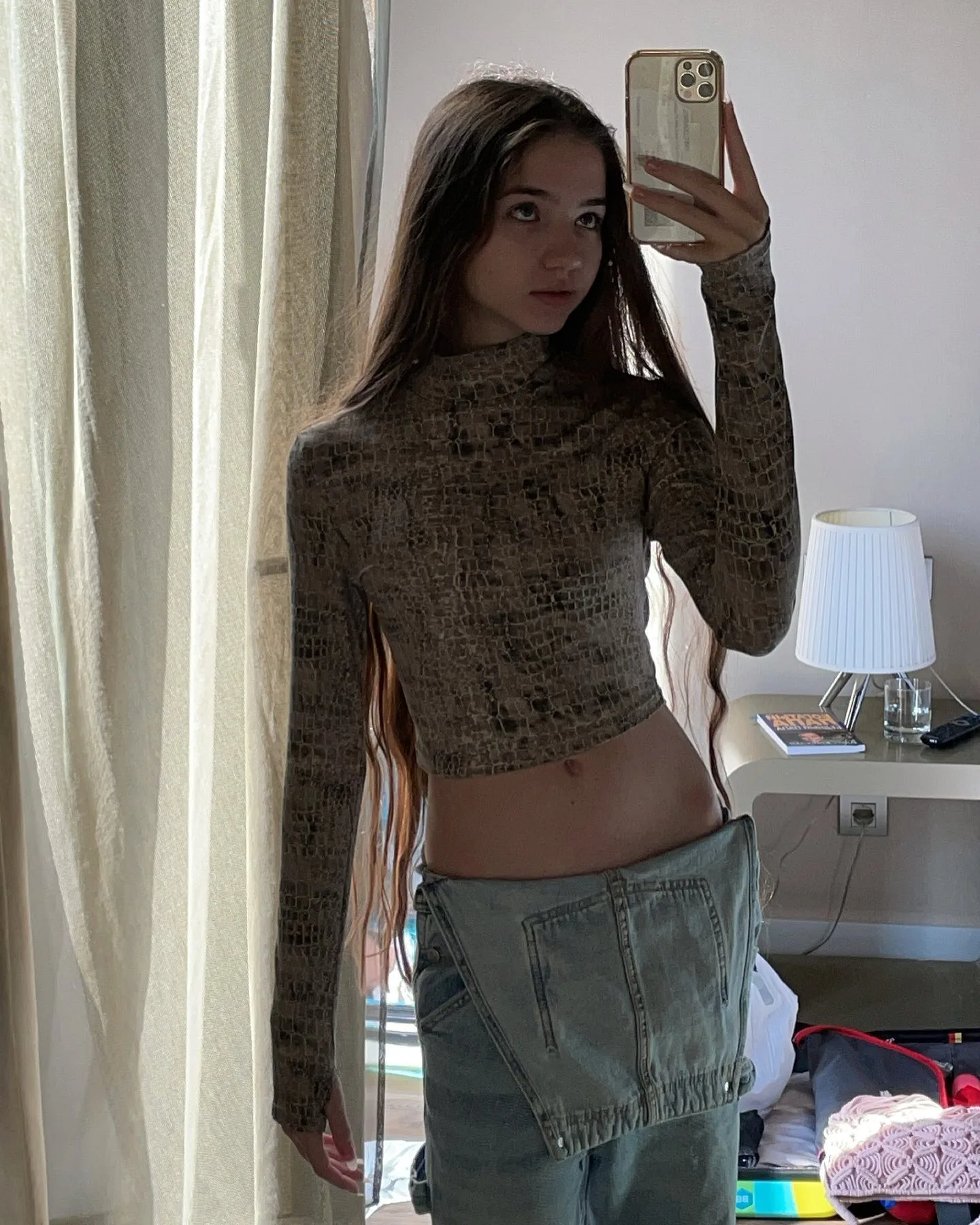 A young woman with long brown hair takes a mirror selfie wearing a brown patterned long-sleeve crop top and light blue jeans with a denim apron skirt.