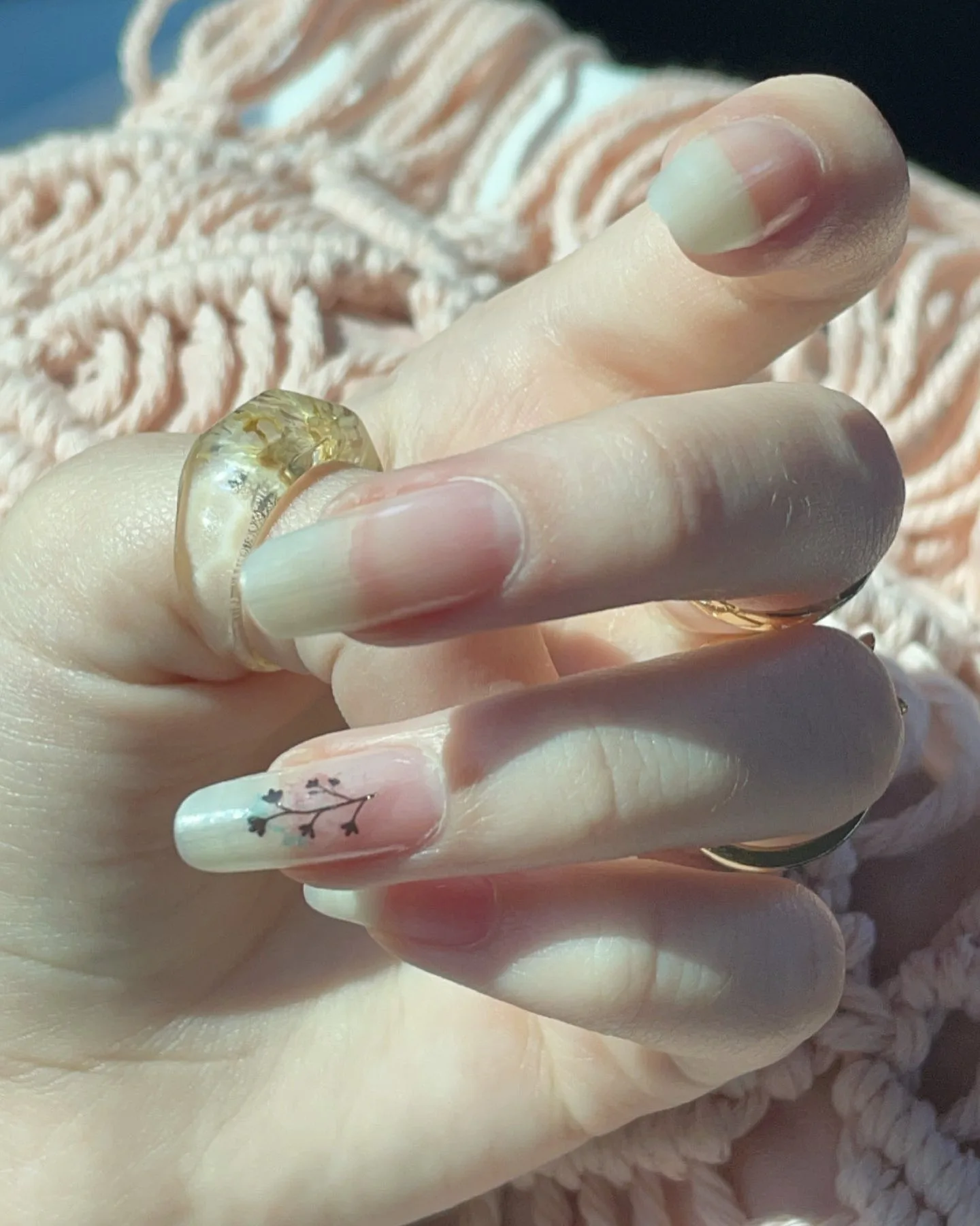Close-up of a hand with natural nails, featuring a white tip with a small black flower design and a gold resin ring, resting on a pink knitted sweater.