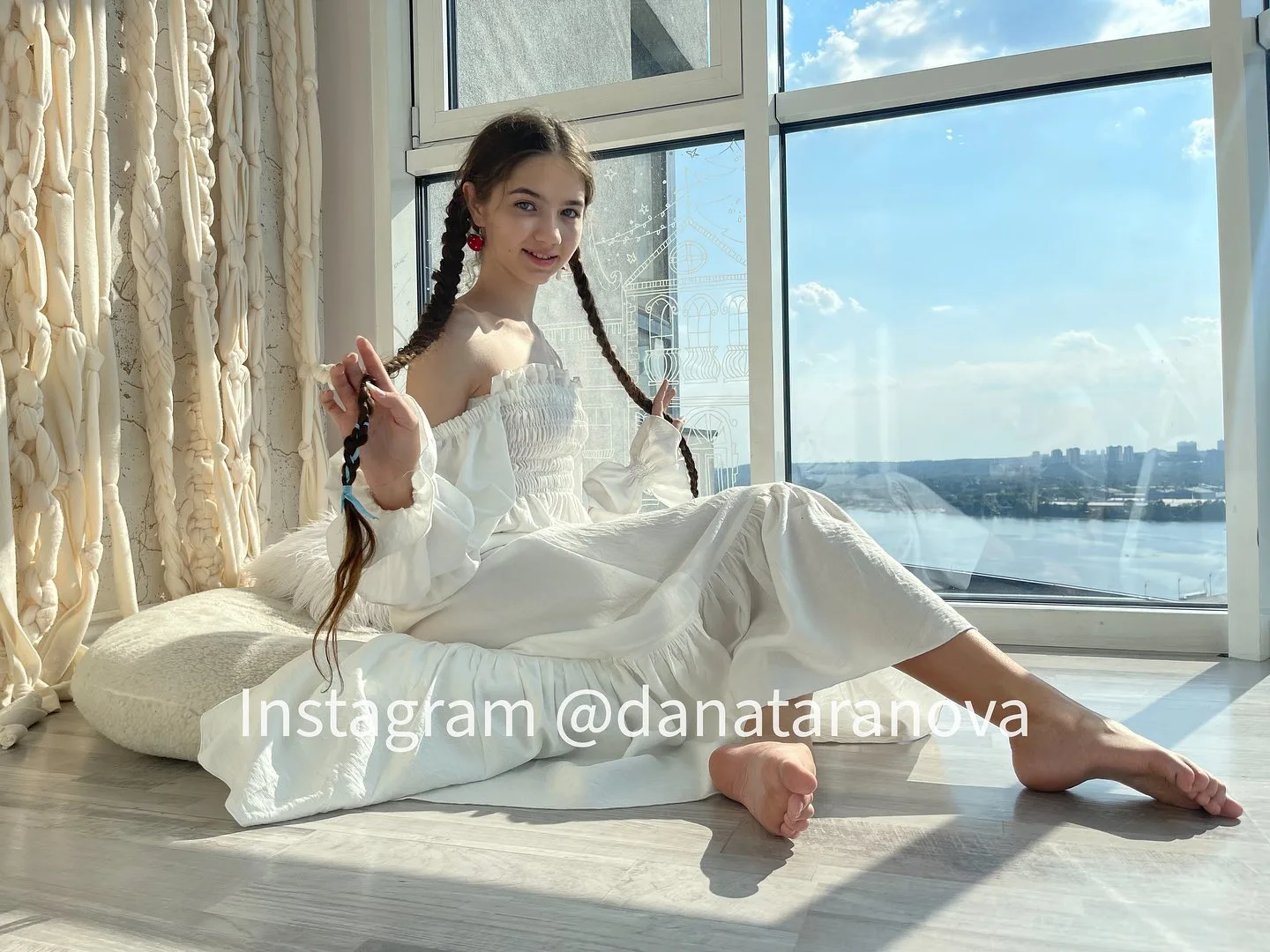 Young woman with braided hair sitting on a white cushion in a white outfit near a large window with a city view.