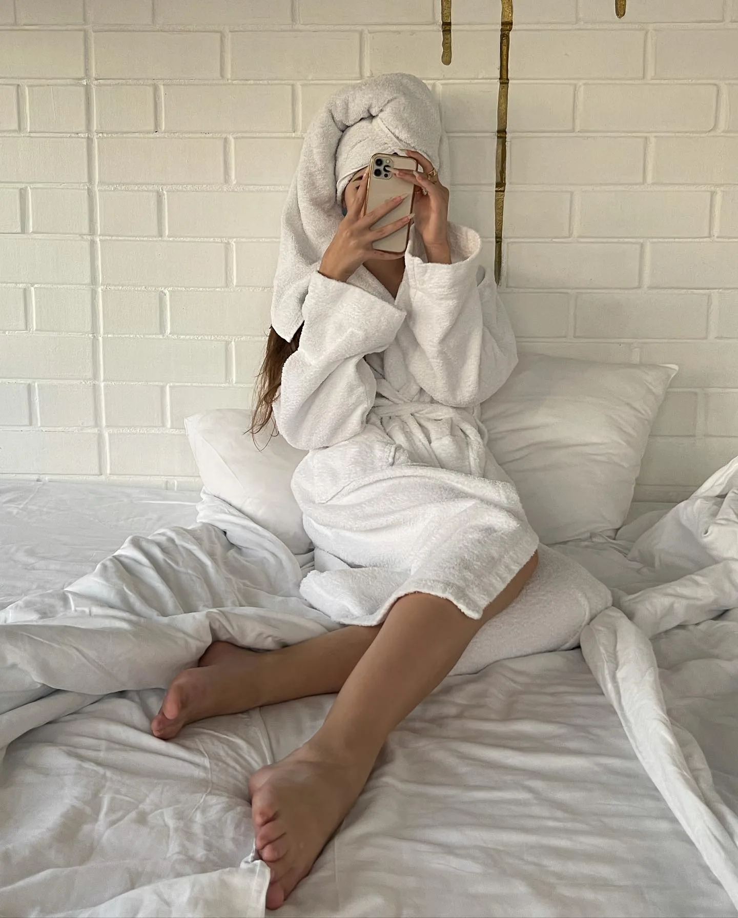 Woman in white bathrobe and towel on head taking a mirror selfie in bed with white linens and brick wall background.