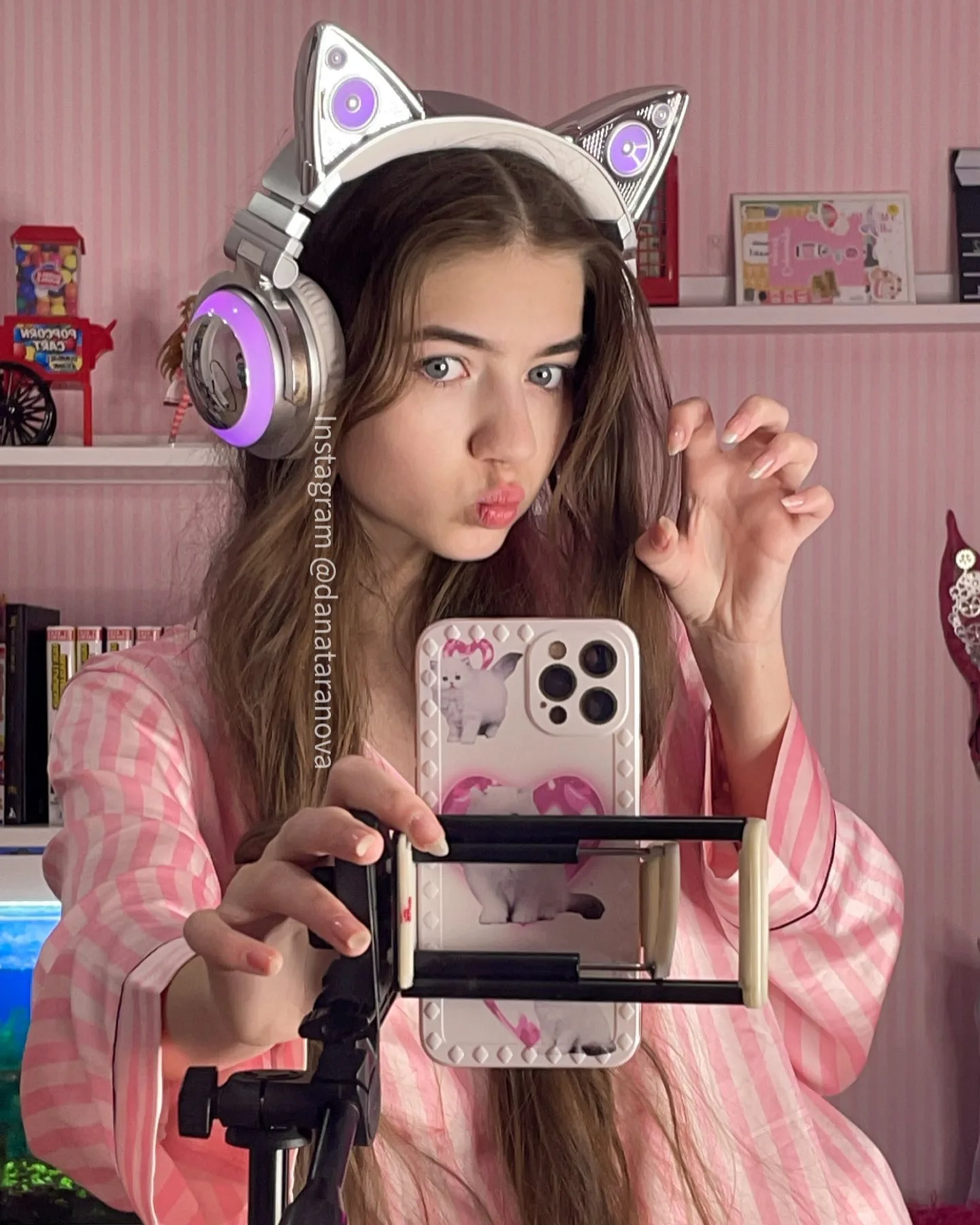 A young woman wearing cat ear headphones and pink pajamas takes a mirror selfie with her phone mounted on a tripod.