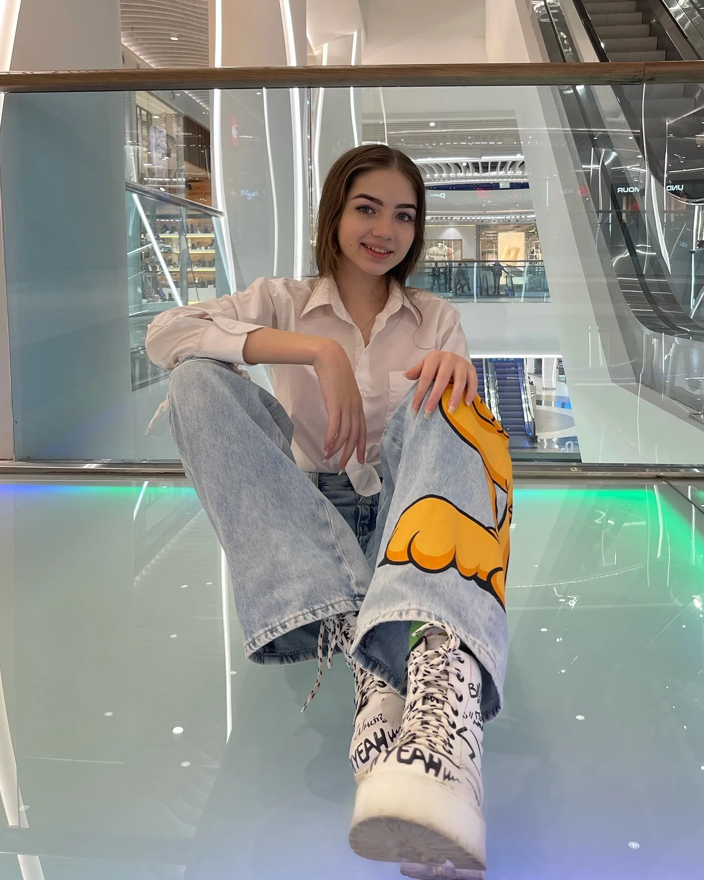 Dana sitting on a glass floor in a mall wearing a pink shirt, light blue jeans with a yellow graphic, and leopard print boots.