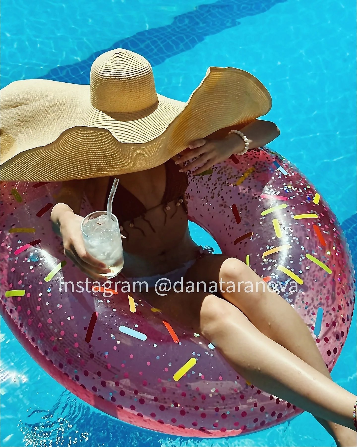 A woman wearing a wide-brimmed straw hat relaxes on a pink donut-shaped pool float with colorful sprinkles in a blue swimming pool, holding a drink.