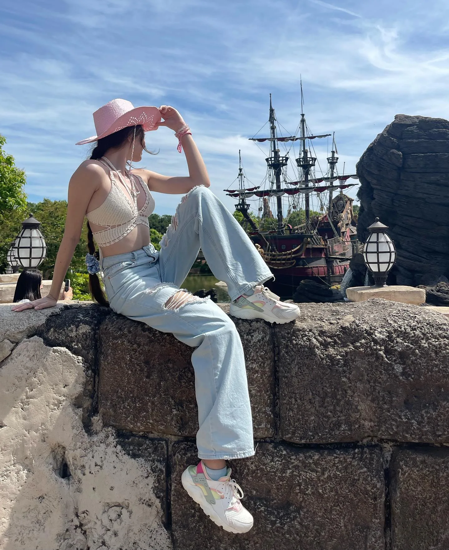 Dana wearing a pink hat and light blue jeans sits on a stone wall with a pirate ship in the background.