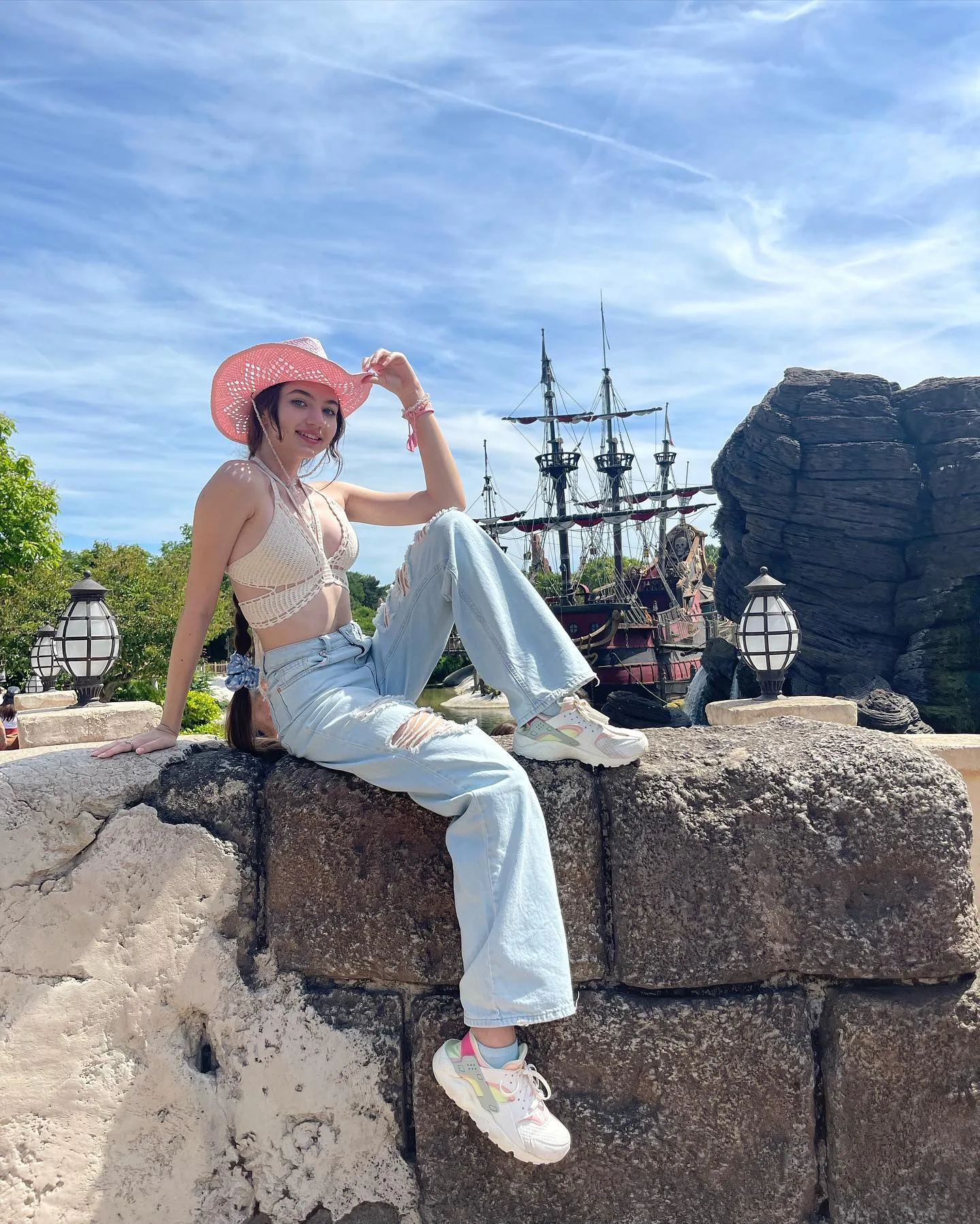 Dana wearing a pink hat and light blue jeans sits on a stone wall with a pirate ship in the background.