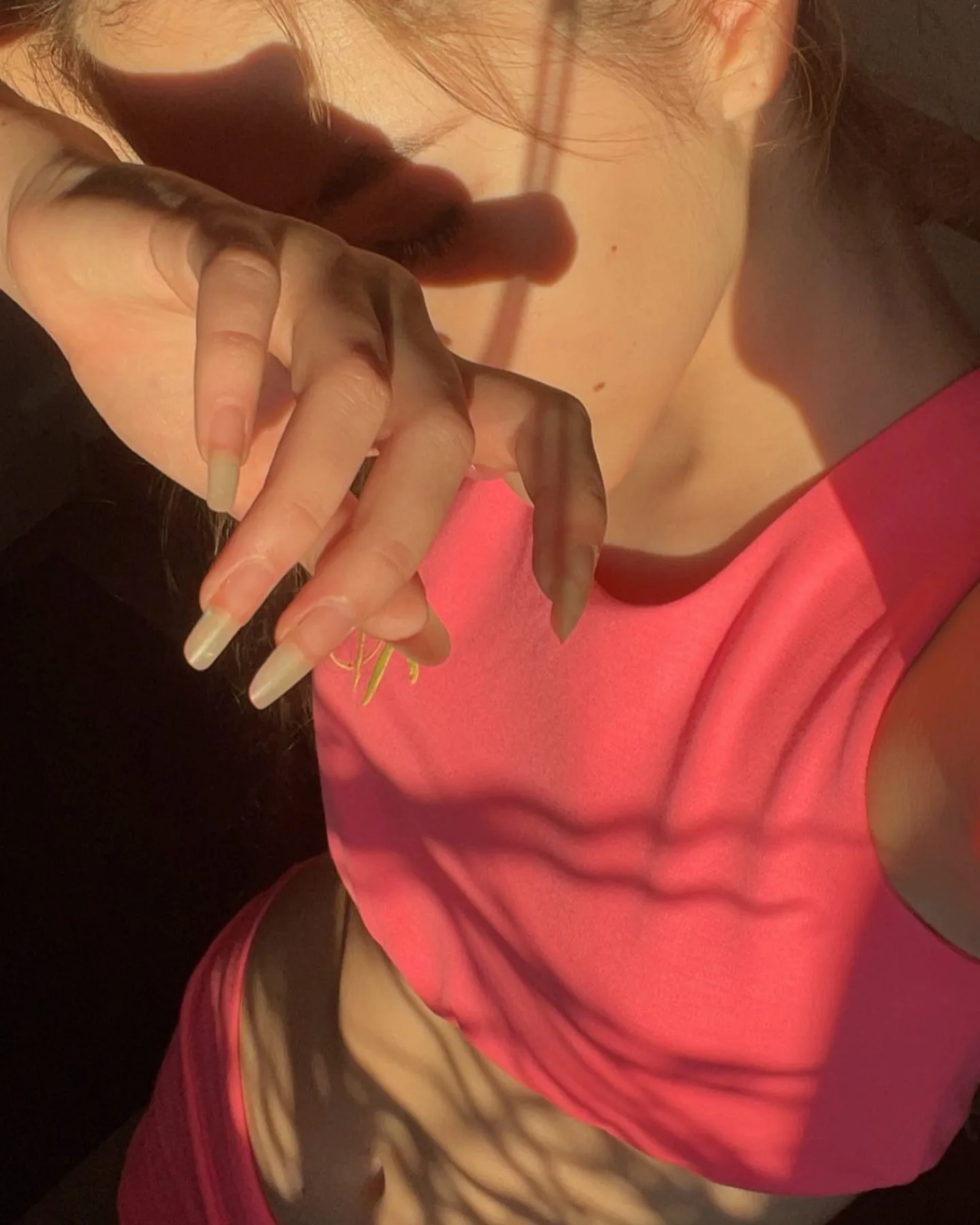 Close-up of a woman in a pink top with long nails, illuminated by warm sunlight casting shadows across her face and hand.