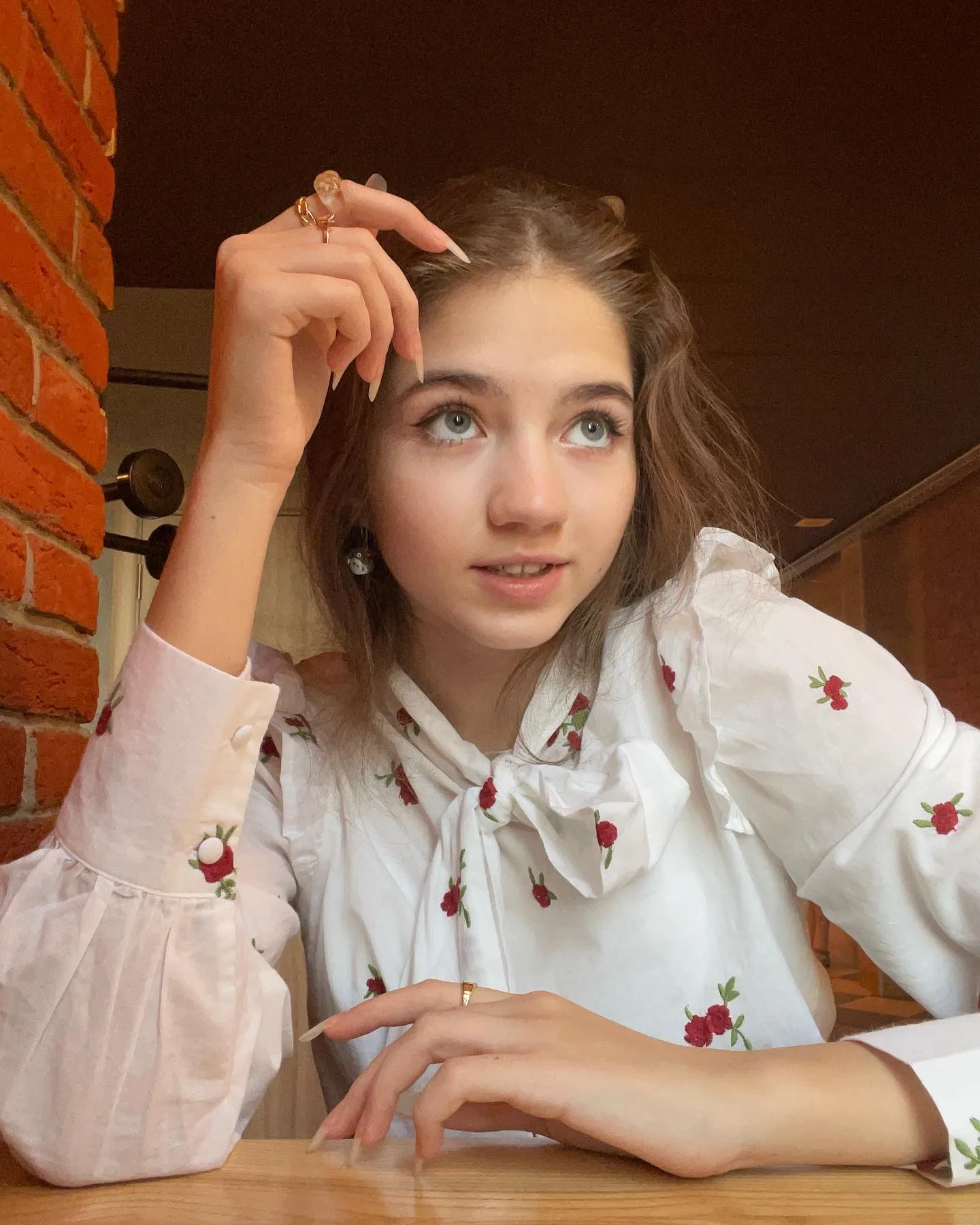 Young woman with brown hair wearing a white blouse with red floral embroidery and a bow tie, posing with one hand near her head against a brick wall background.