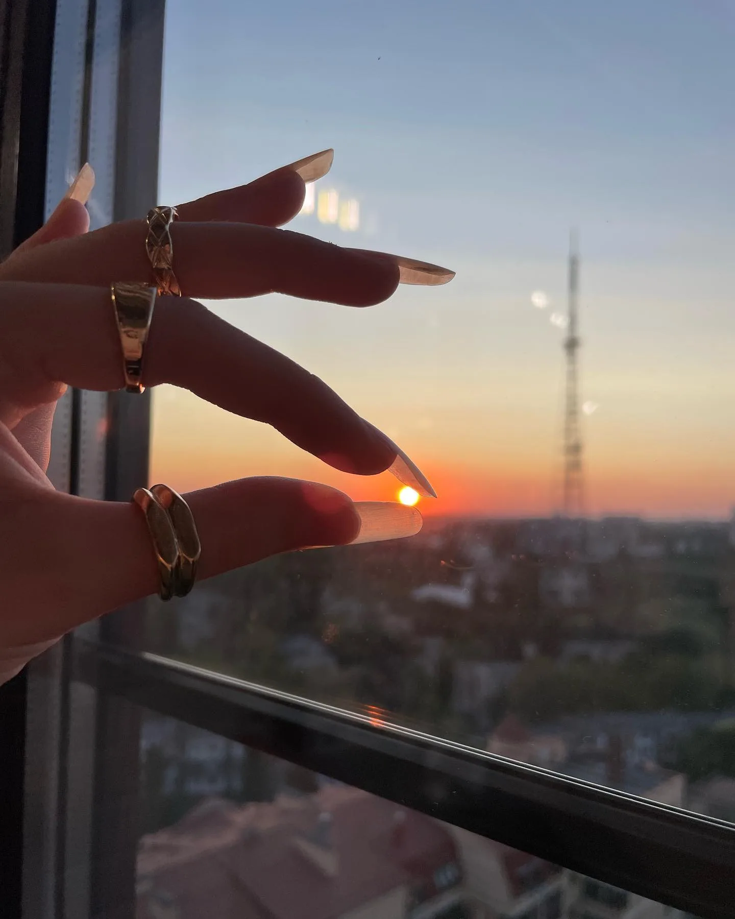 A hand with long nails and gold rings frames the setting sun through a window, with a cityscape and tower visible in the background.