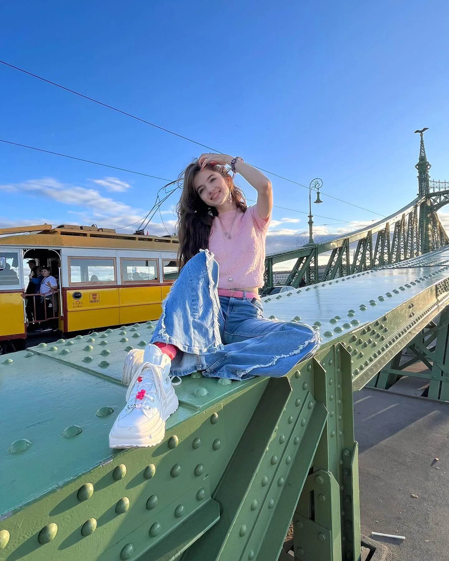 A woman in a pink shirt and blue jeans sits on a green bridge railing with a yellow tram in the background.