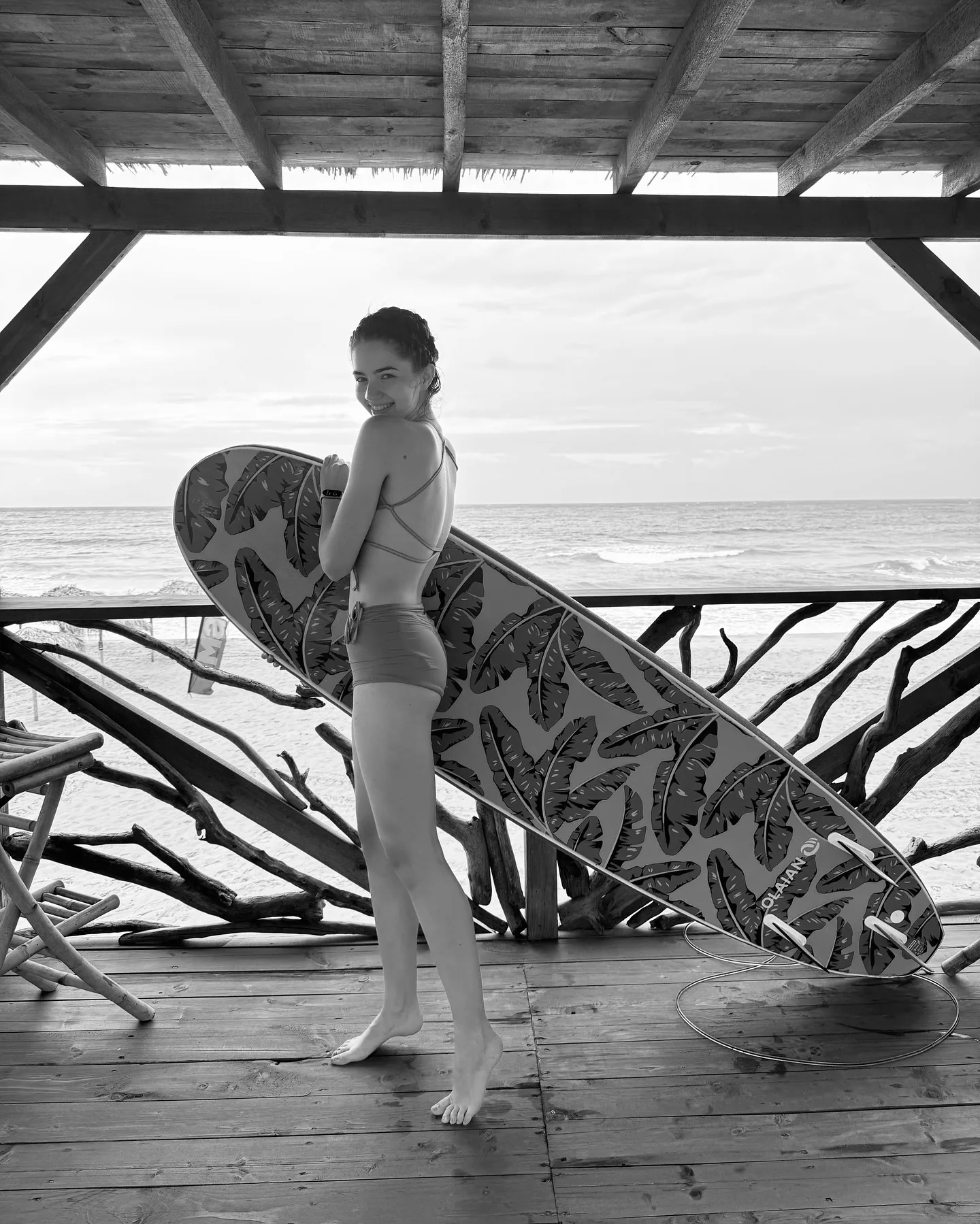 A woman in a bikini stands on a wooden deck holding a surfboard with a leaf pattern, looking back at the camera with the ocean in the background.