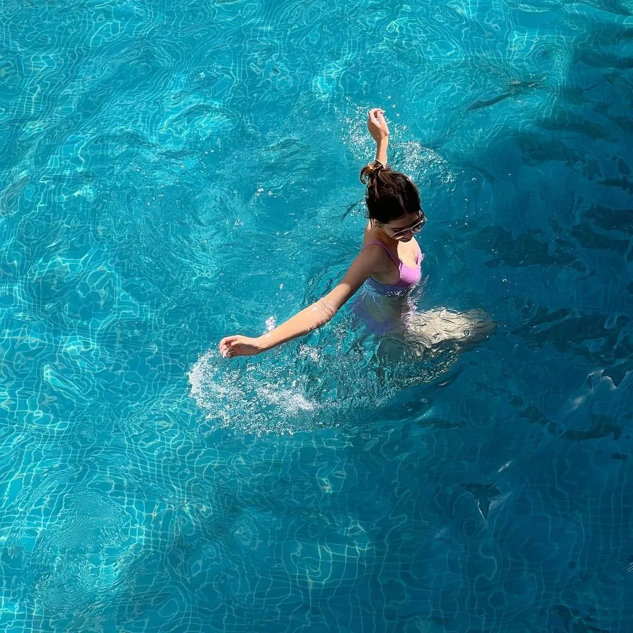 A woman in a pink swimsuit splashing in a blue swimming pool, viewed from above.