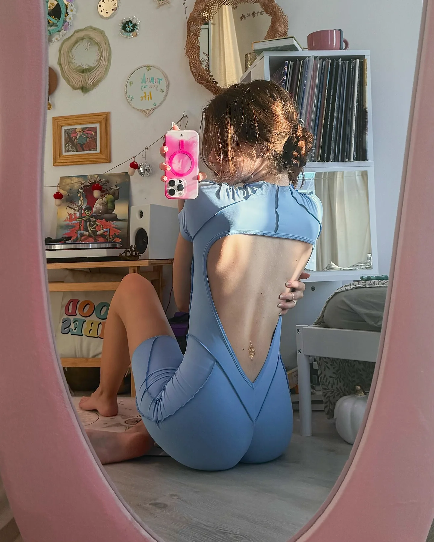 A woman in a blue cutout bodysuit takes a mirror selfie in a pink-framed mirror, showing her back and a cozy room with vinyl records.