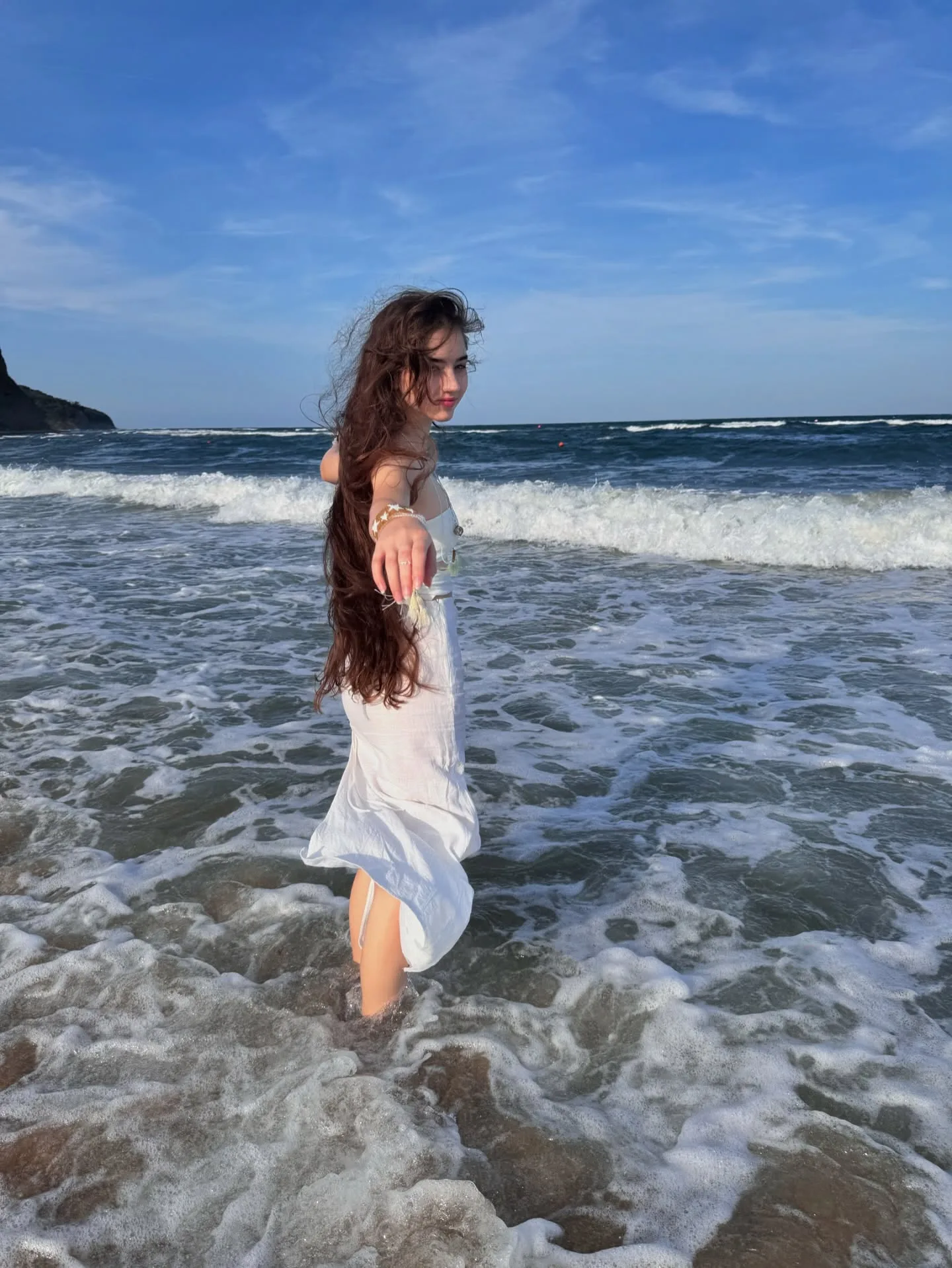 A woman in a white dress stands in shallow ocean waves with long brown hair blowing in the wind against a blue sky.