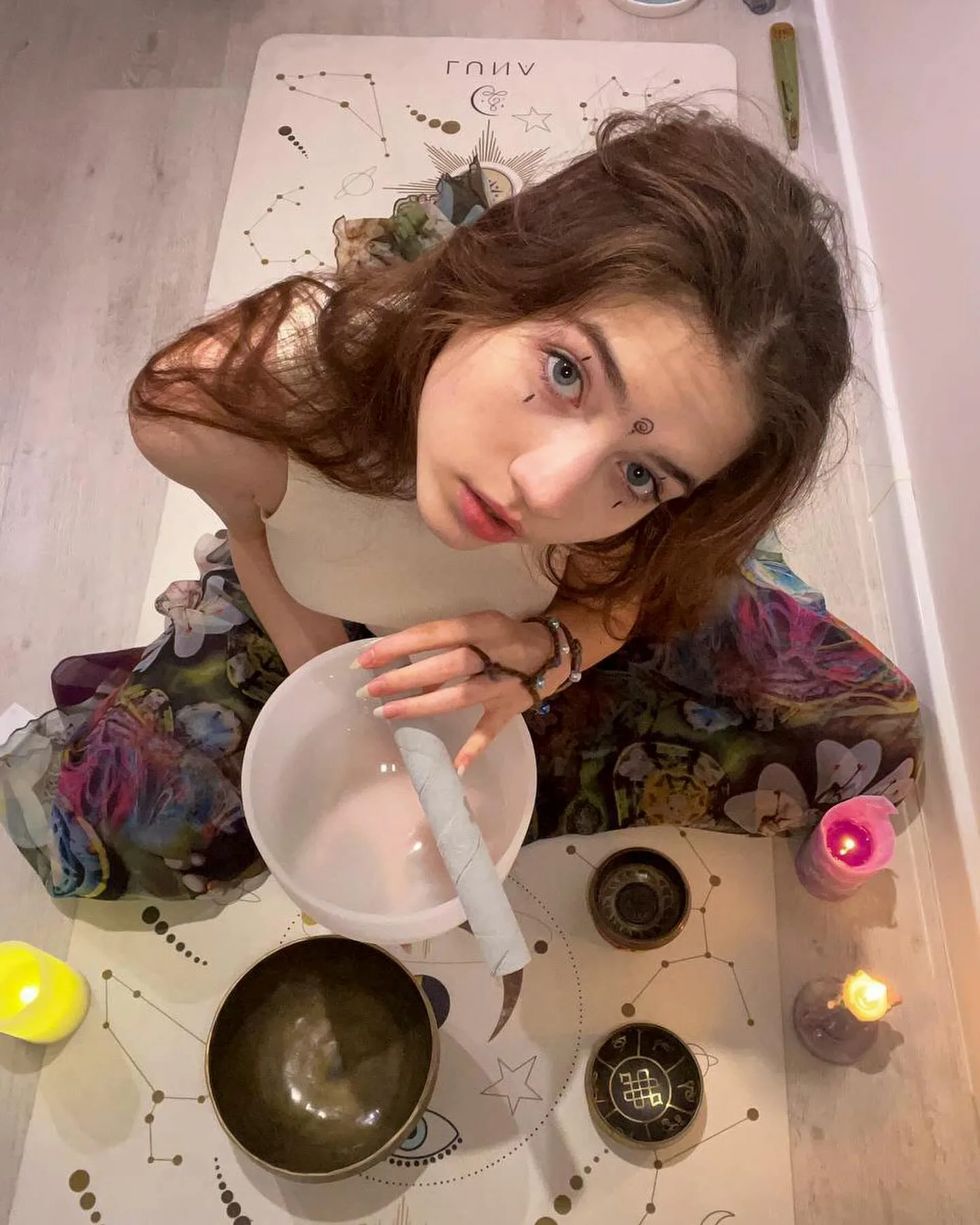 Dana sitting on a star chart rug with singing bowls, candles, and a bowl, looking up at the camera.