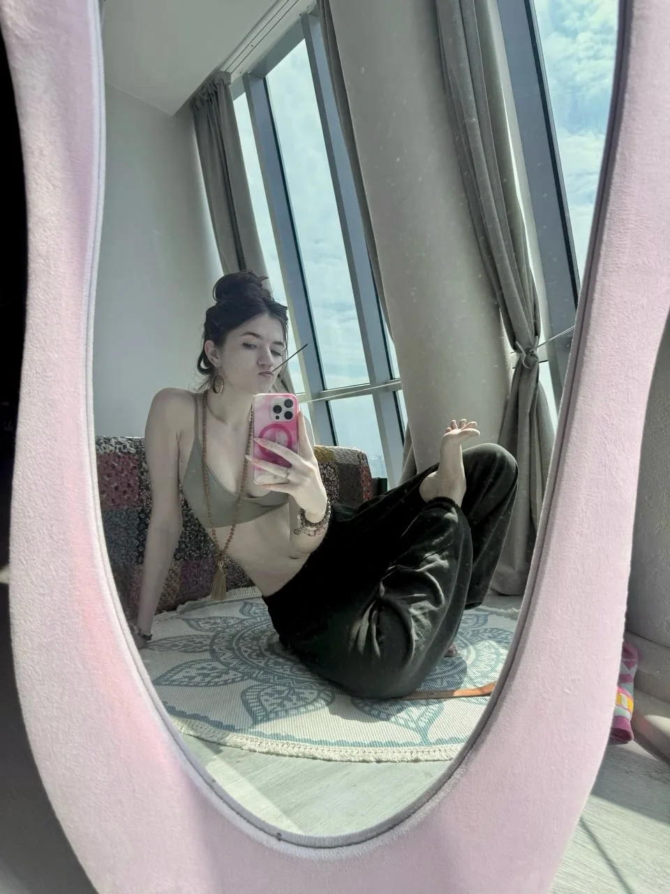 Woman taking a mirror selfie in a pink oval mirror, wearing a beige top and black leather pants, sitting on a patterned rug near a window.