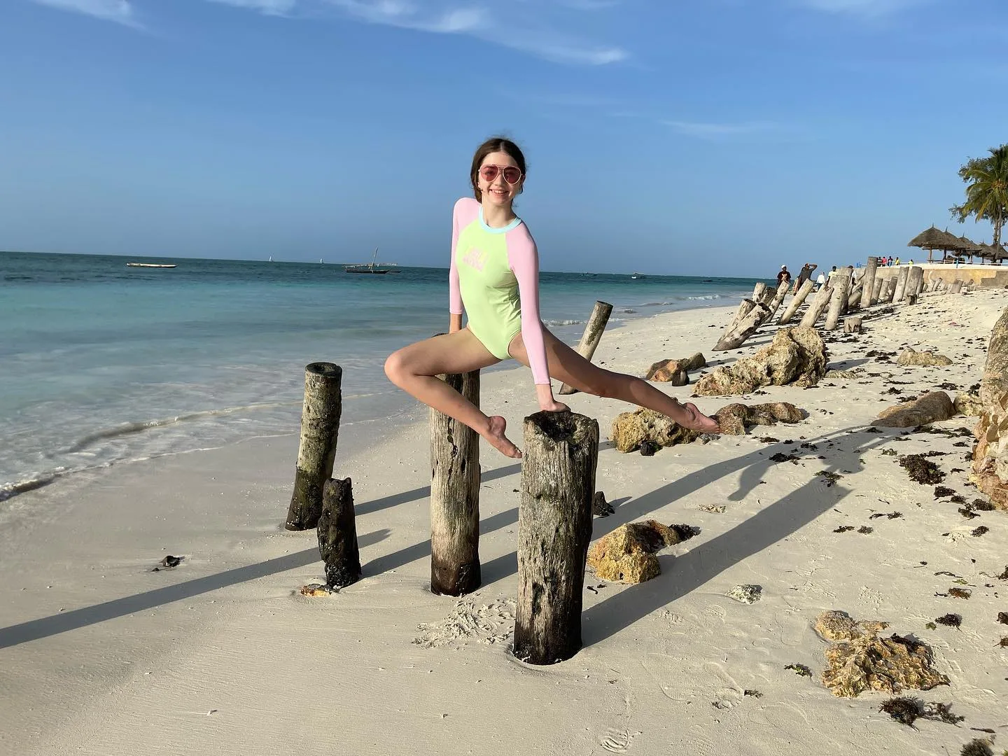 A woman in a neon green and pink swimsuit poses on wooden posts on a sandy beach with turquoise water and a clear blue sky.