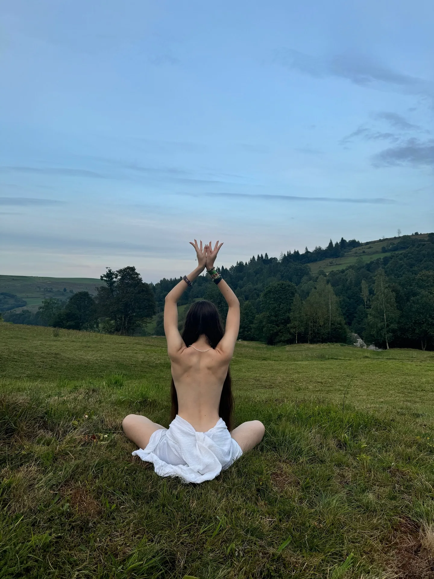 A woman with long dark hair sits cross-legged on a grassy hillside, facing away from the camera with her arms raised in a prayer pose against a backdrop of rolling green hills and 
