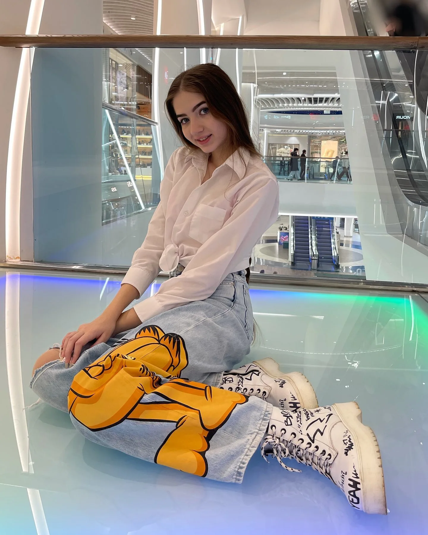Dana sitting on a glowing platform in a mall, wearing a white shirt, light blue jeans with a large yellow cartoon graphic, and white high-top sneakers.