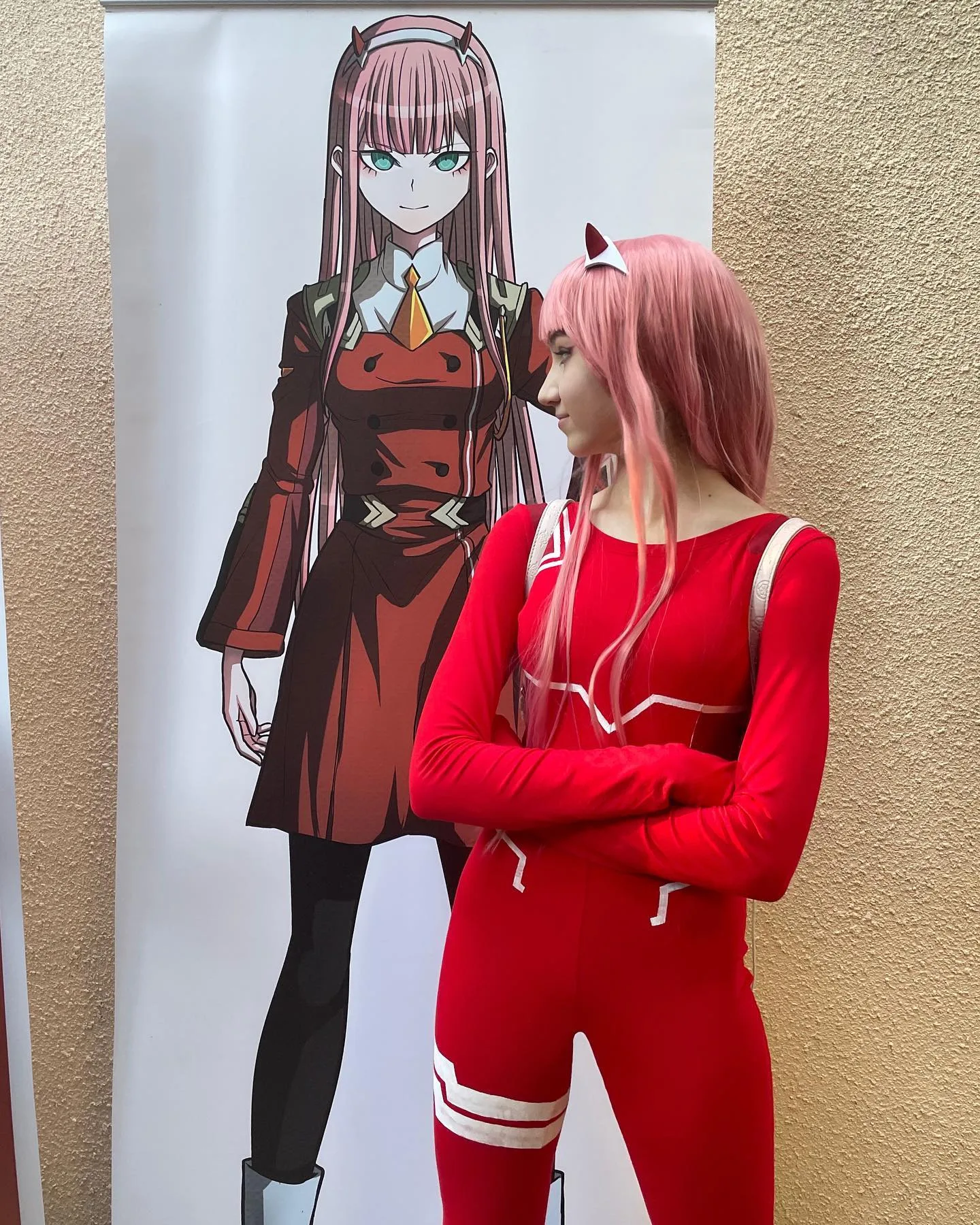A cosplayer dressed as Zero Two from Darling in the Franxx stands with arms crossed next to a large poster of the anime character.