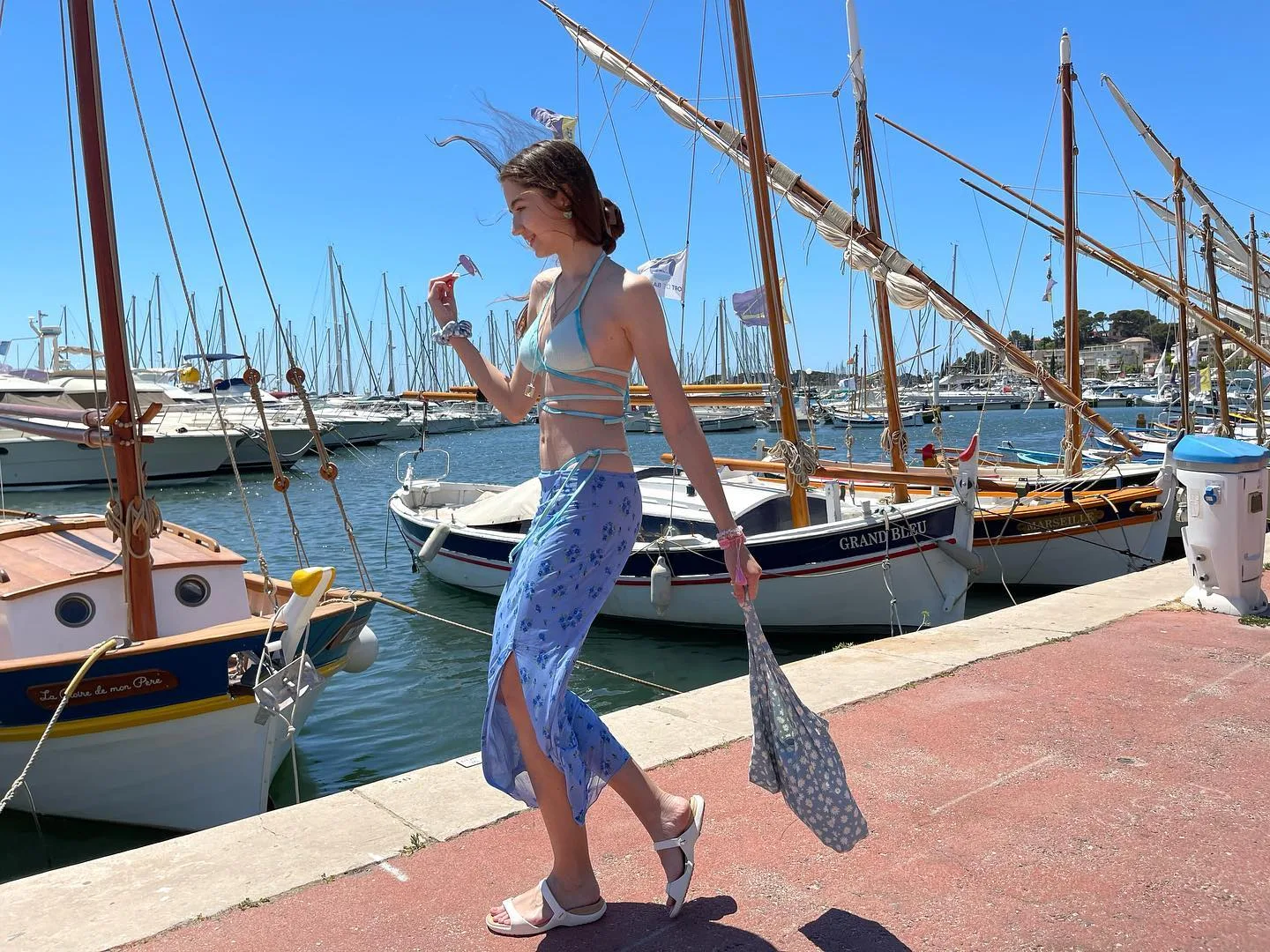 Woman in blue floral skirt walking on a sunny harbor pier with sailboats in the background.