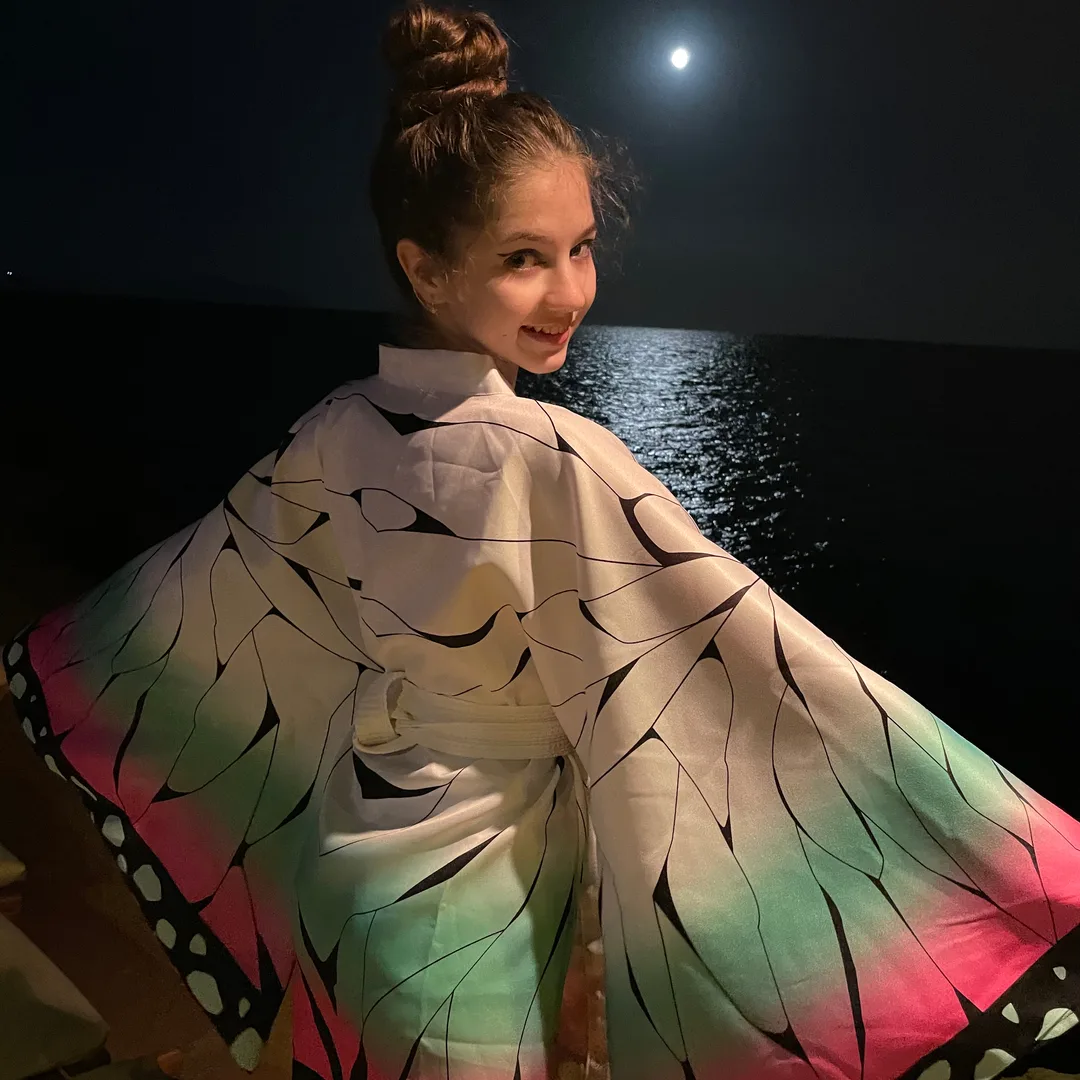 Woman in a colorful butterfly wing haori posing by the water at night with a full moon in the background.