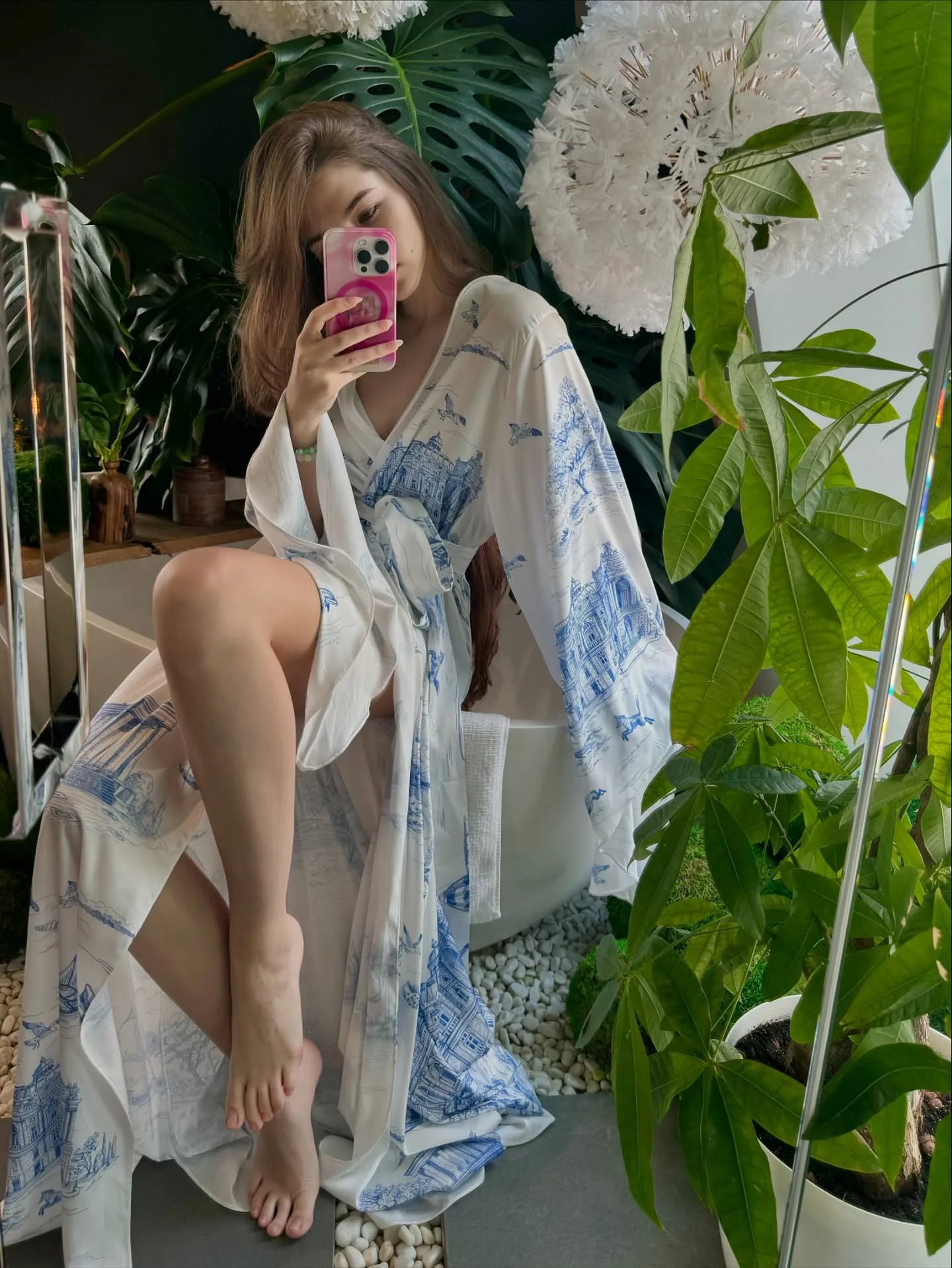 A woman in a white and blue floral robe takes a mirror selfie while sitting on a white chair surrounded by large green plants and white flowers.