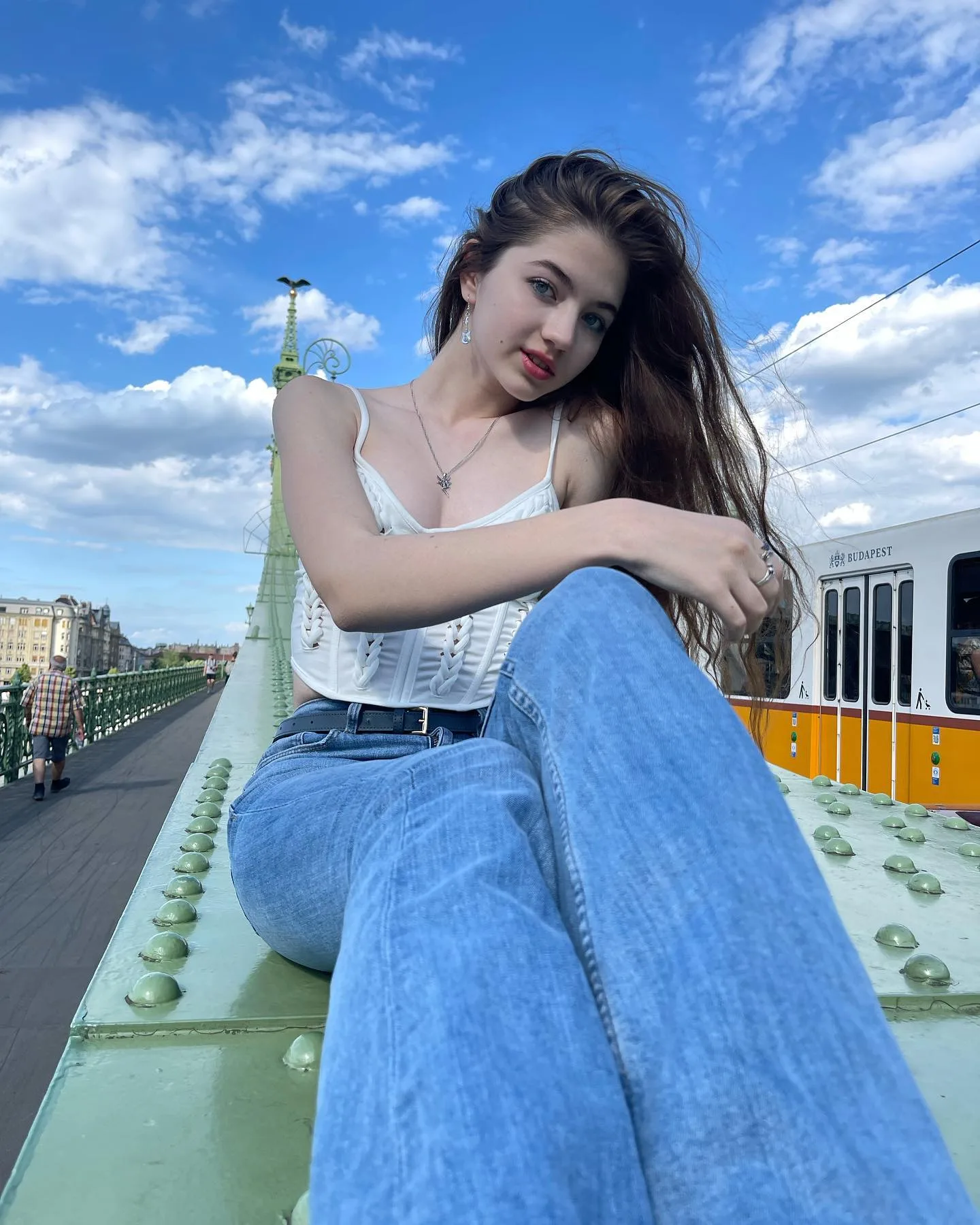Dana sitting on a green bridge railing wearing a white top and blue jeans against a blue sky.
