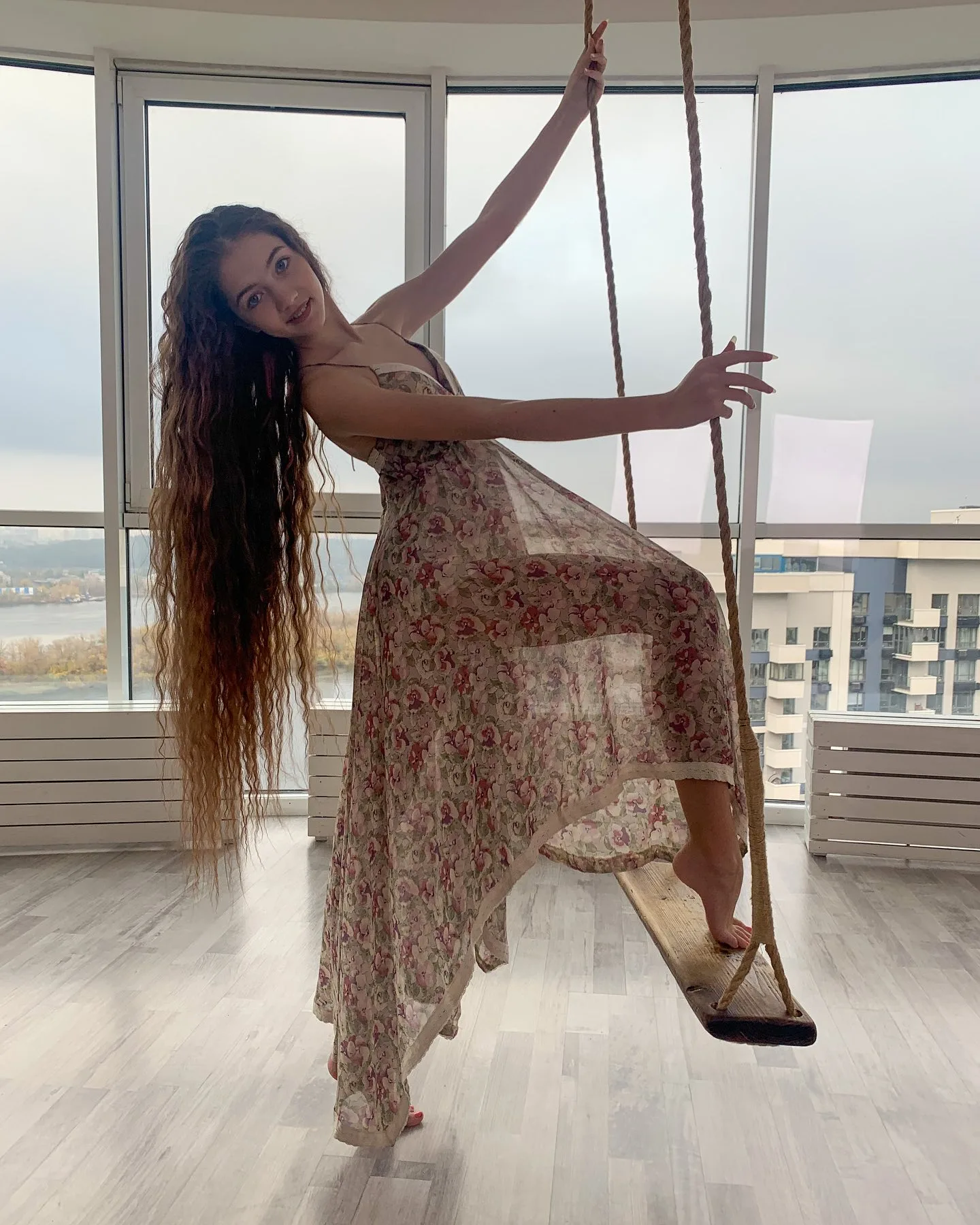 Dana posing on a wooden swing in a floral dress inside a bright room with large windows.