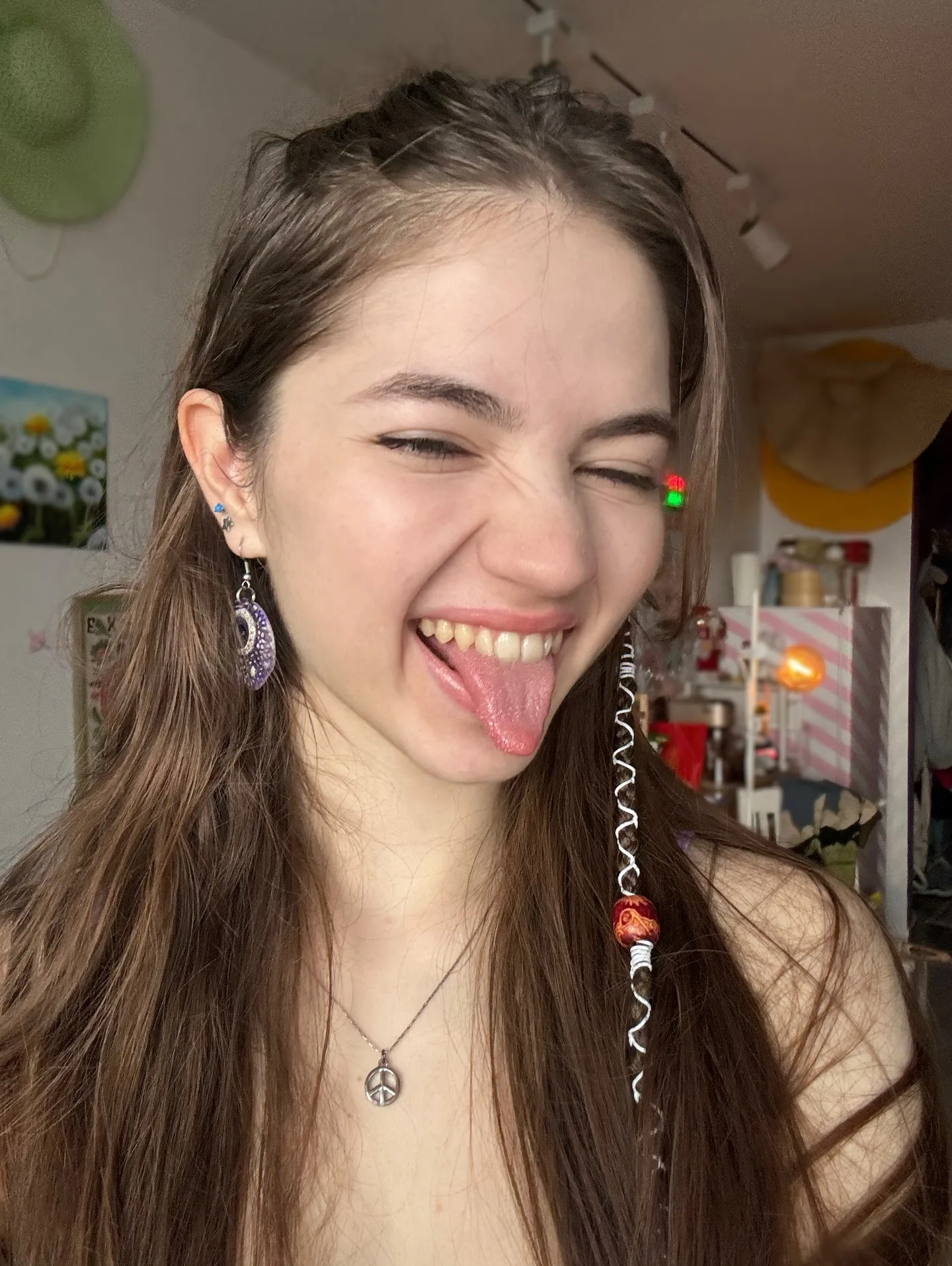 A young woman with long brown hair winks and sticks out her tongue, wearing a necklace and earrings in a room with hats on the wall.