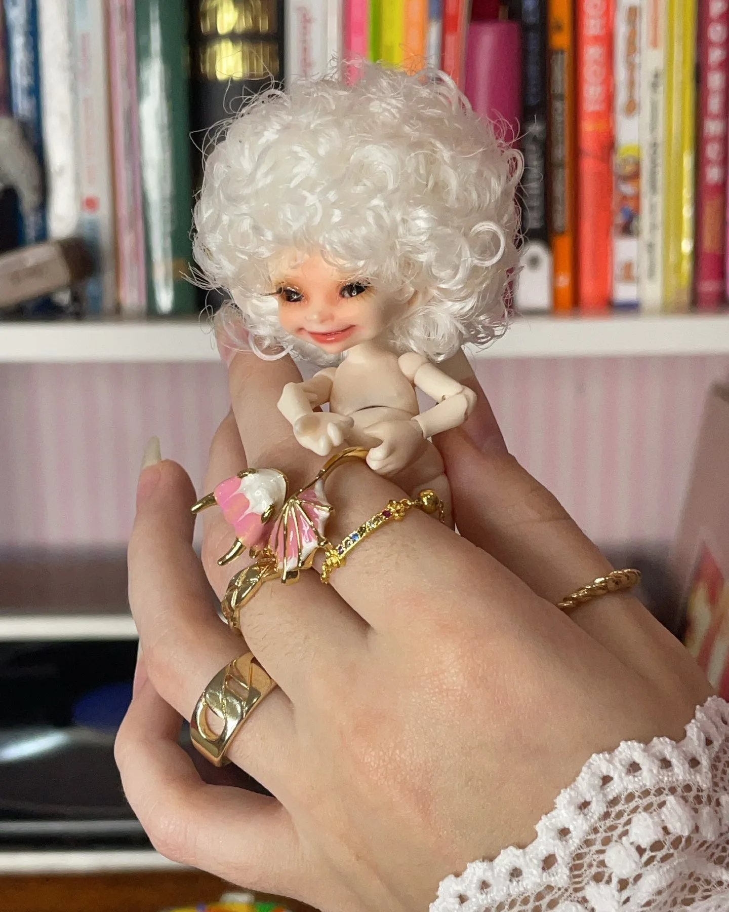 A hand adorned with multiple gold rings holds a small doll with curly white hair and a pink headband against a blurred bookshelf background.