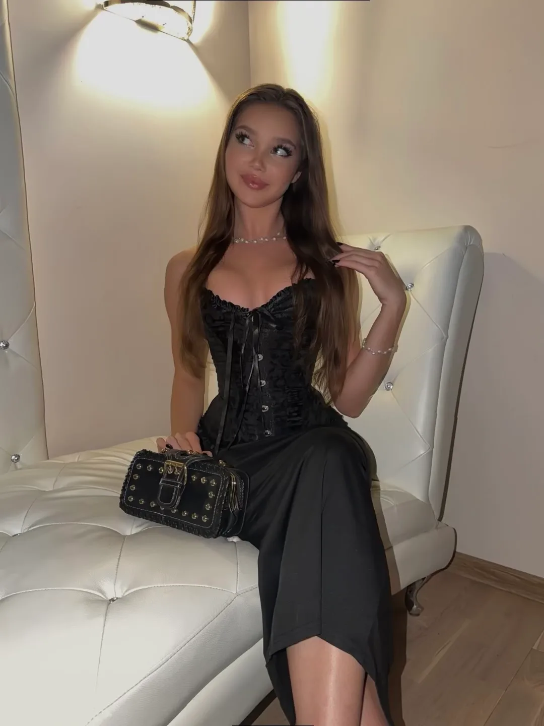 Tina Abramova posing in a black corset dress and holding a studded handbag while sitting on a white tufted chaise lounge.
