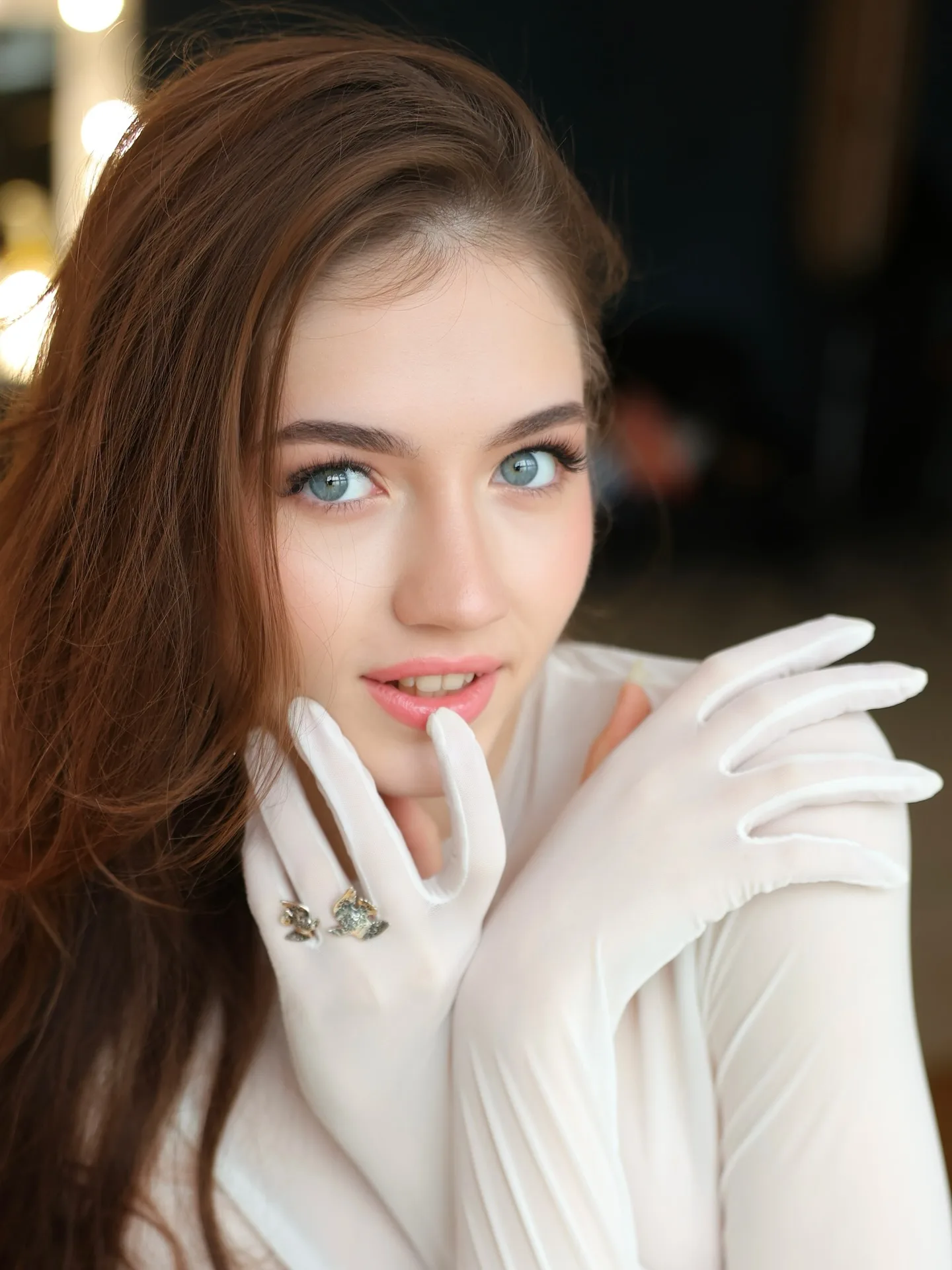 Portrait of a woman with long brown hair and blue eyes wearing white gloves and a white top, posing with her hands near her face.