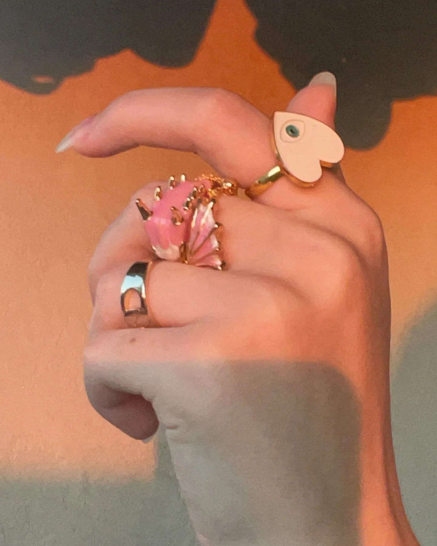 A hand wearing multiple rings including a pink shell ring, a gold band, and a white heart-shaped ring against an orange background.