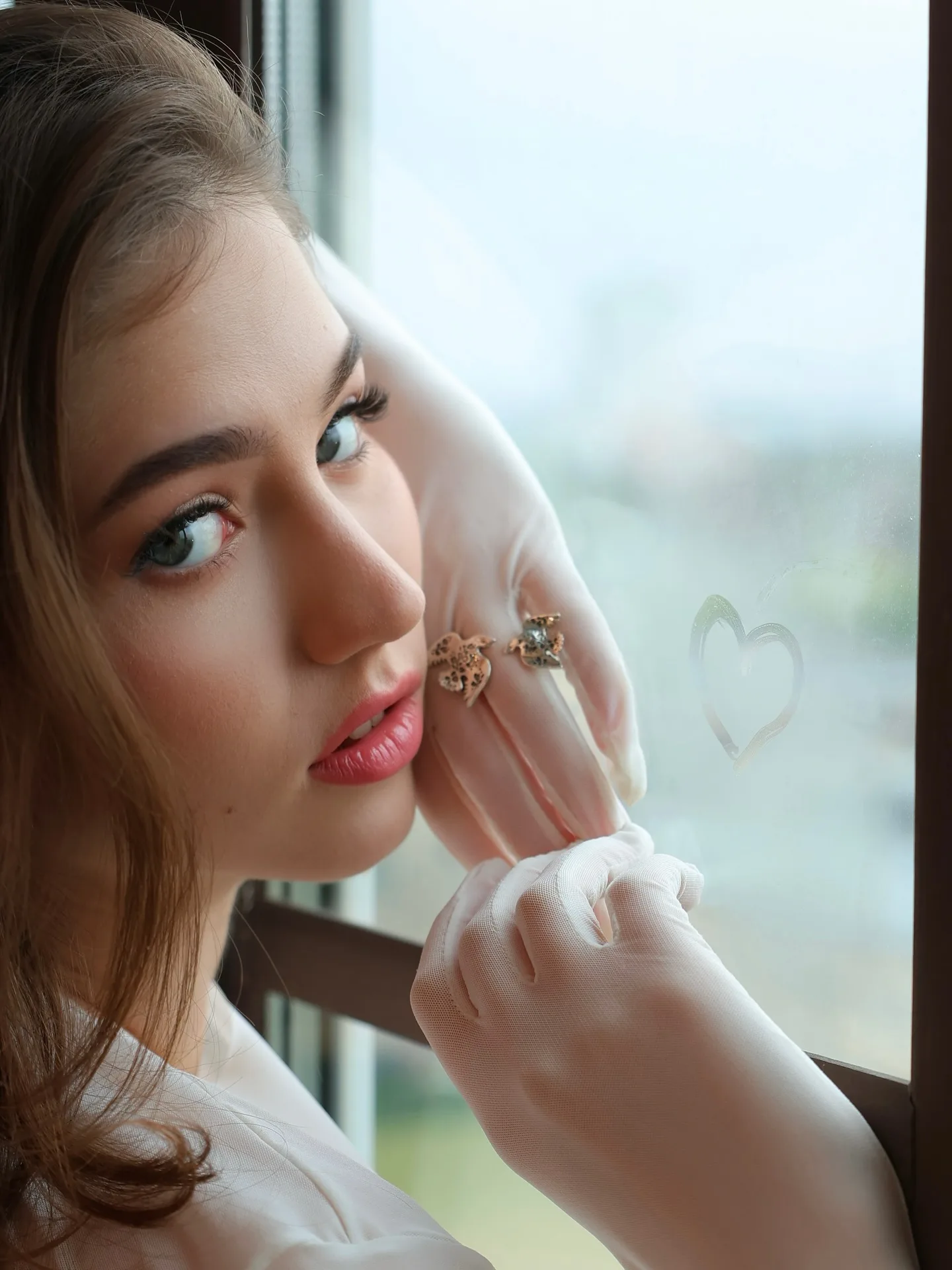 Young woman with brown hair and blue eyes resting her chin on her hand near a window with a heart drawn on the glass.