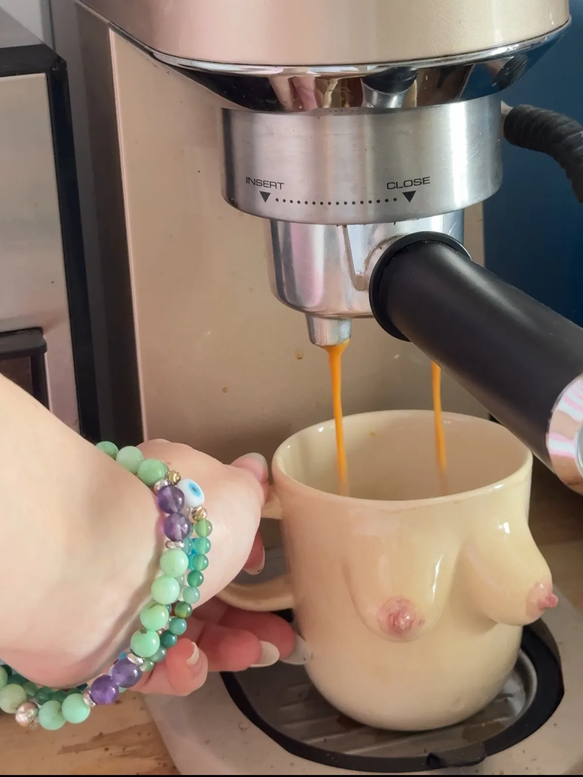 Coffee pouring from an espresso machine into a beige ceramic mug shaped like breasts, held by a hand with a beaded bracelet.
