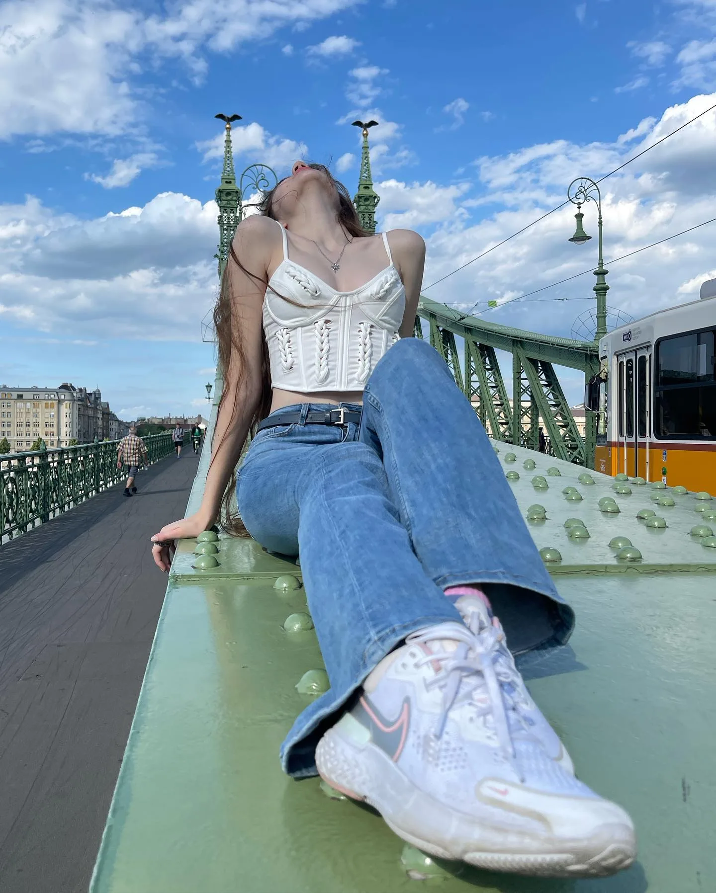 Woman in white corset and blue jeans leaning back on a green bridge railing under a blue sky.