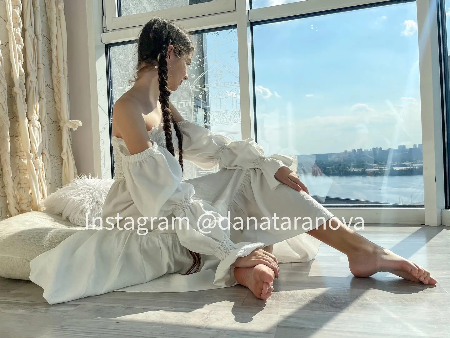 A woman with braided hair sits on a cushion by a large window in a white dress, looking out at a sunny waterfront view.